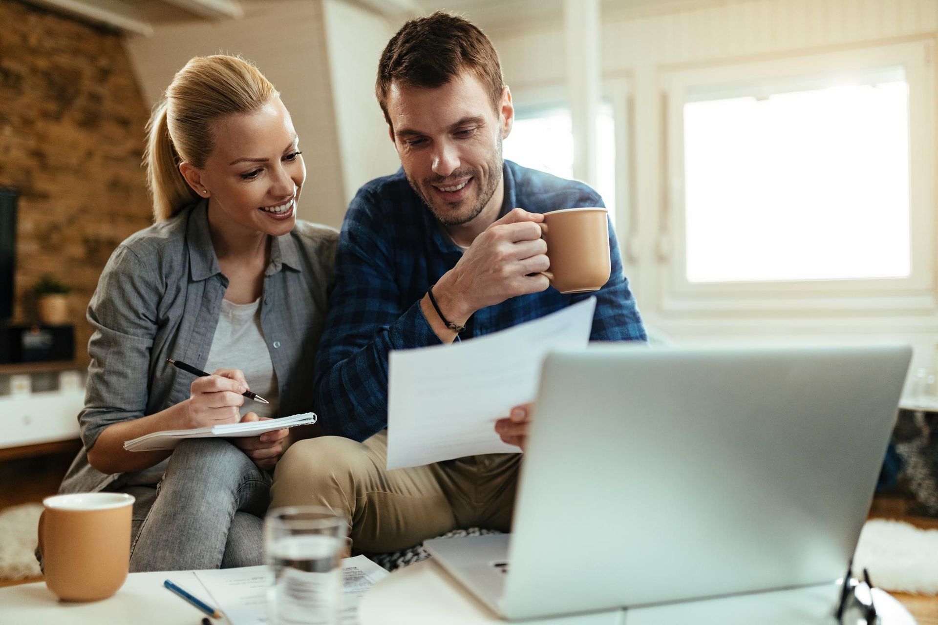A couple sitting together at home, reviewing documents on a laptop and writing notes while holding mugs of coffee.