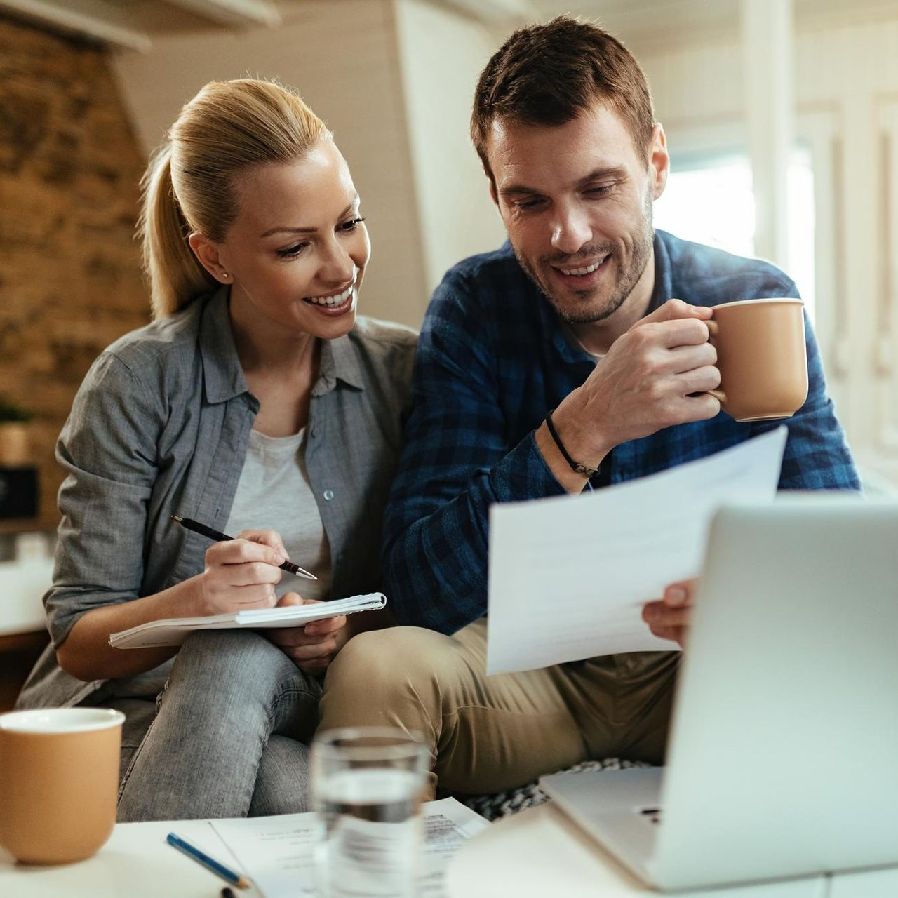 A smiling couple in a home setting reviews documents and a laptop while holding coffee mugs.
