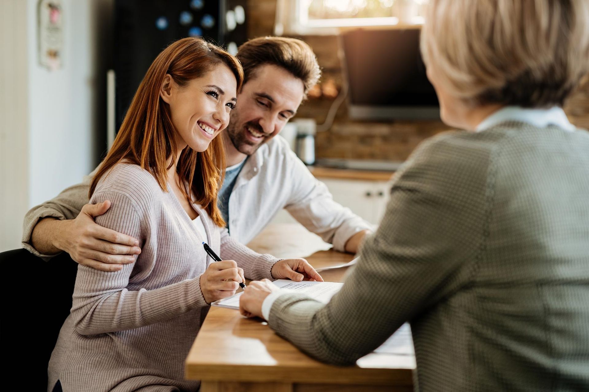 A couple sits at a wooden table smiling as one signs a document while meeting with a professional in an office setting.