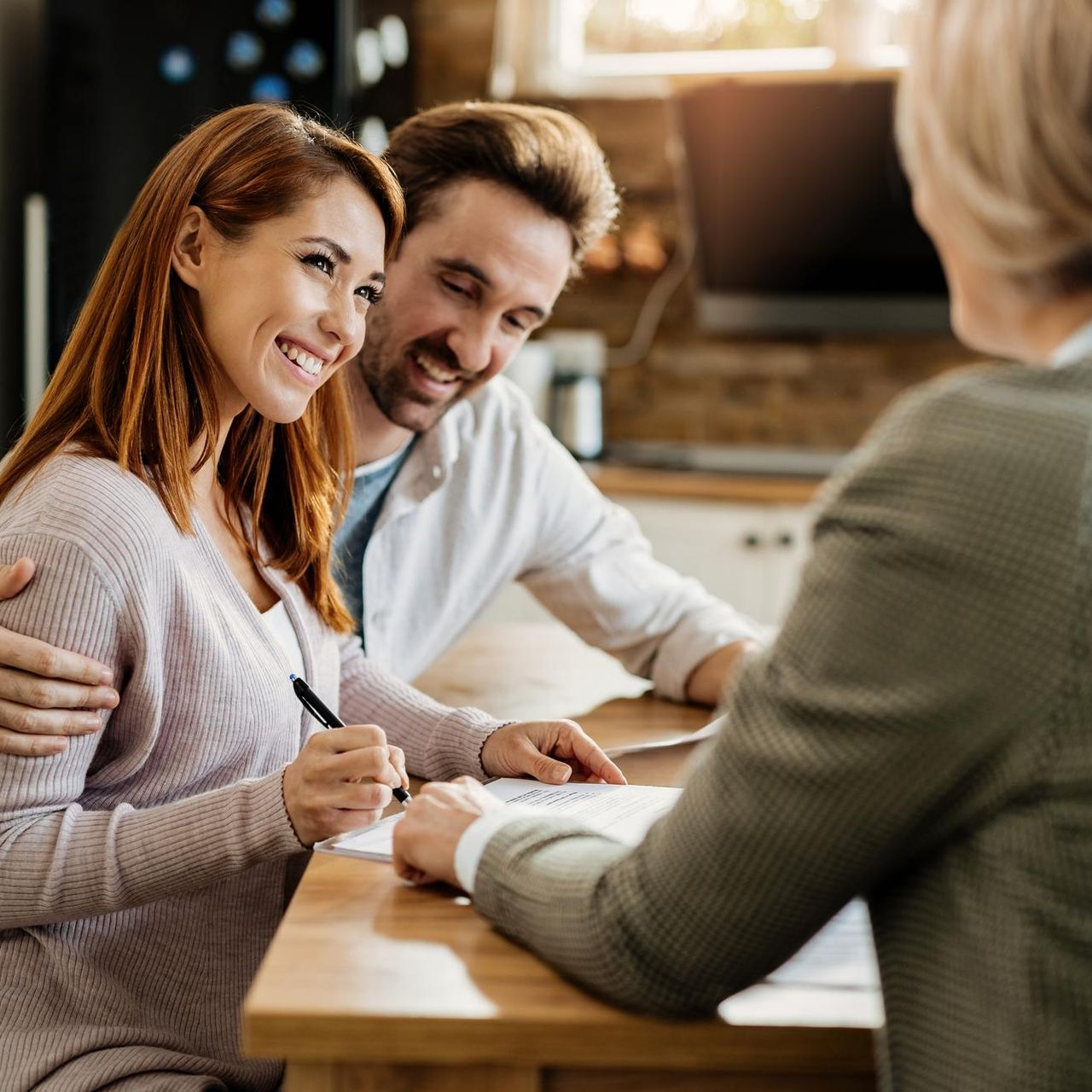 A smiling couple sits at a wooden table signing a document with a professional in a home or office setting.