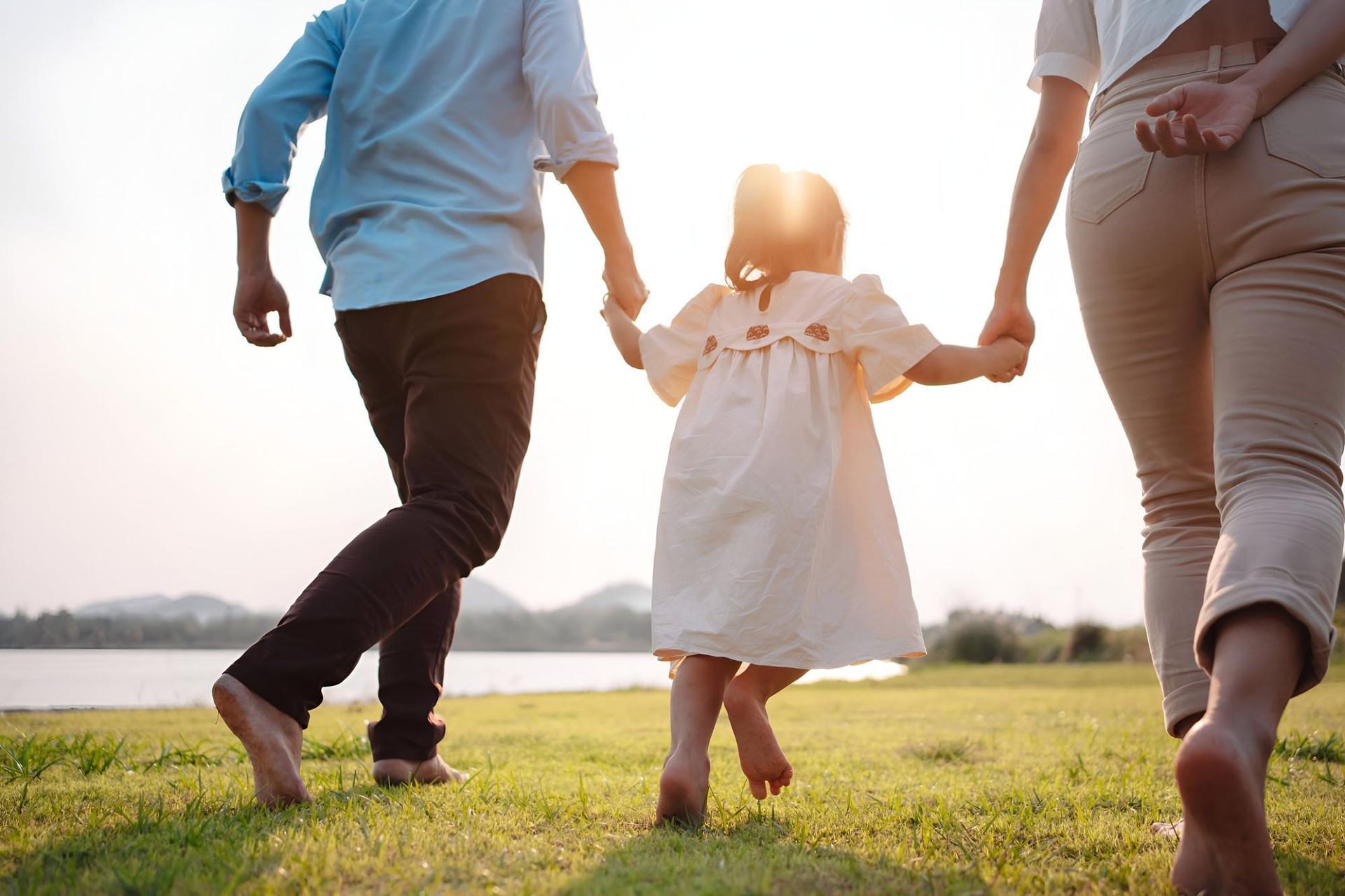 A family holds hands while walking through a grassy field towards a sunset by a lake.