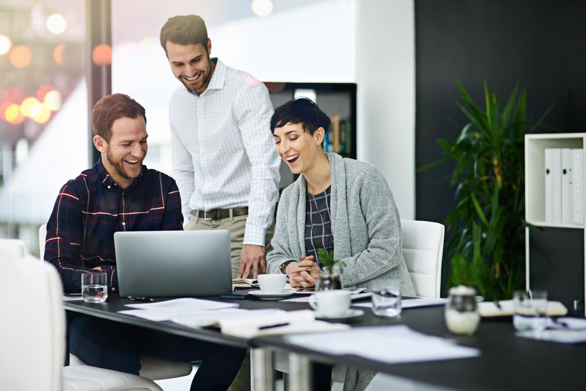 Three coworkers in a bright office laugh while collaborating on a laptop at a table with documents and coffee cups.