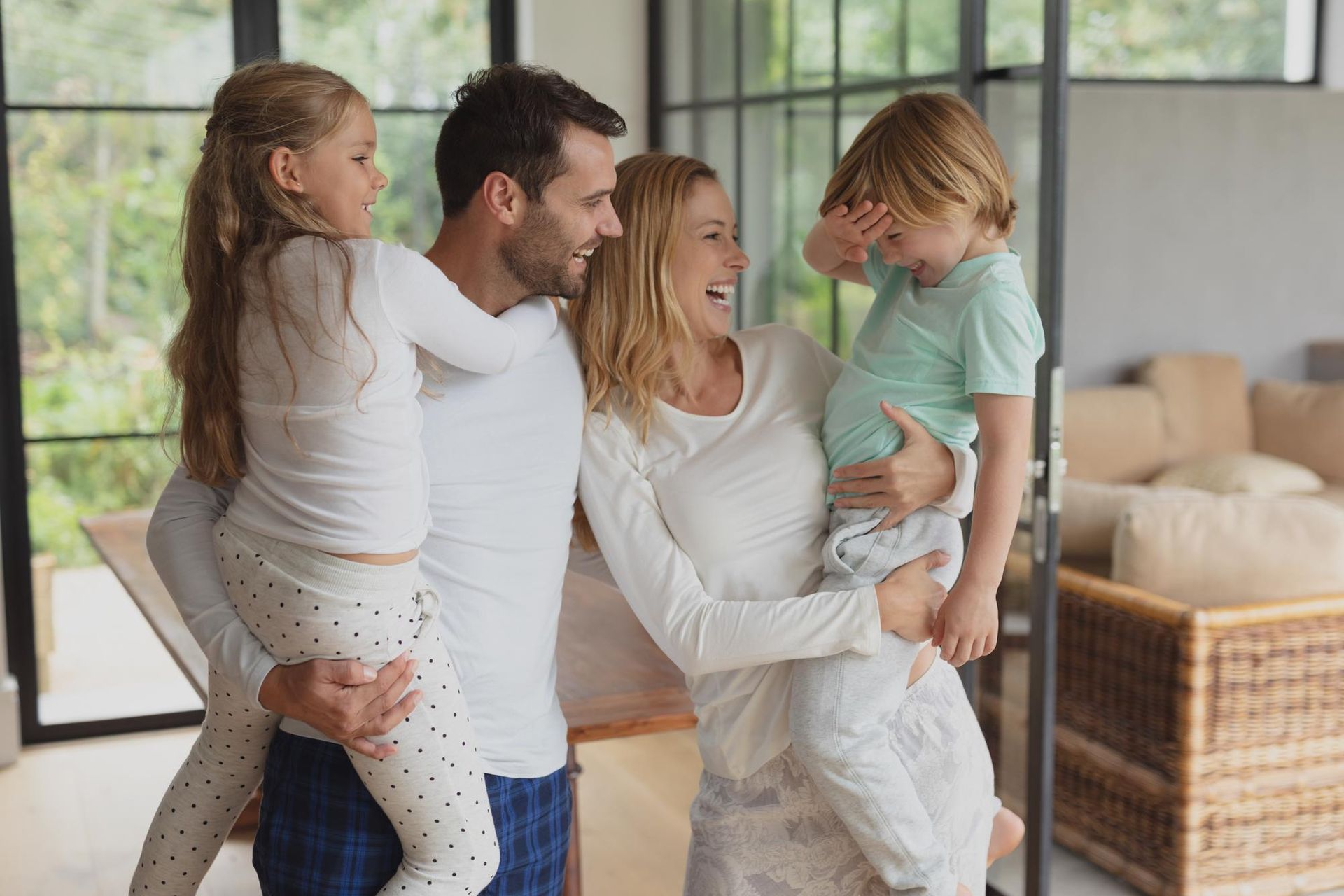 Parents laugh and play in a bright living room, each holding a child while standing in a warm, domestic setting.