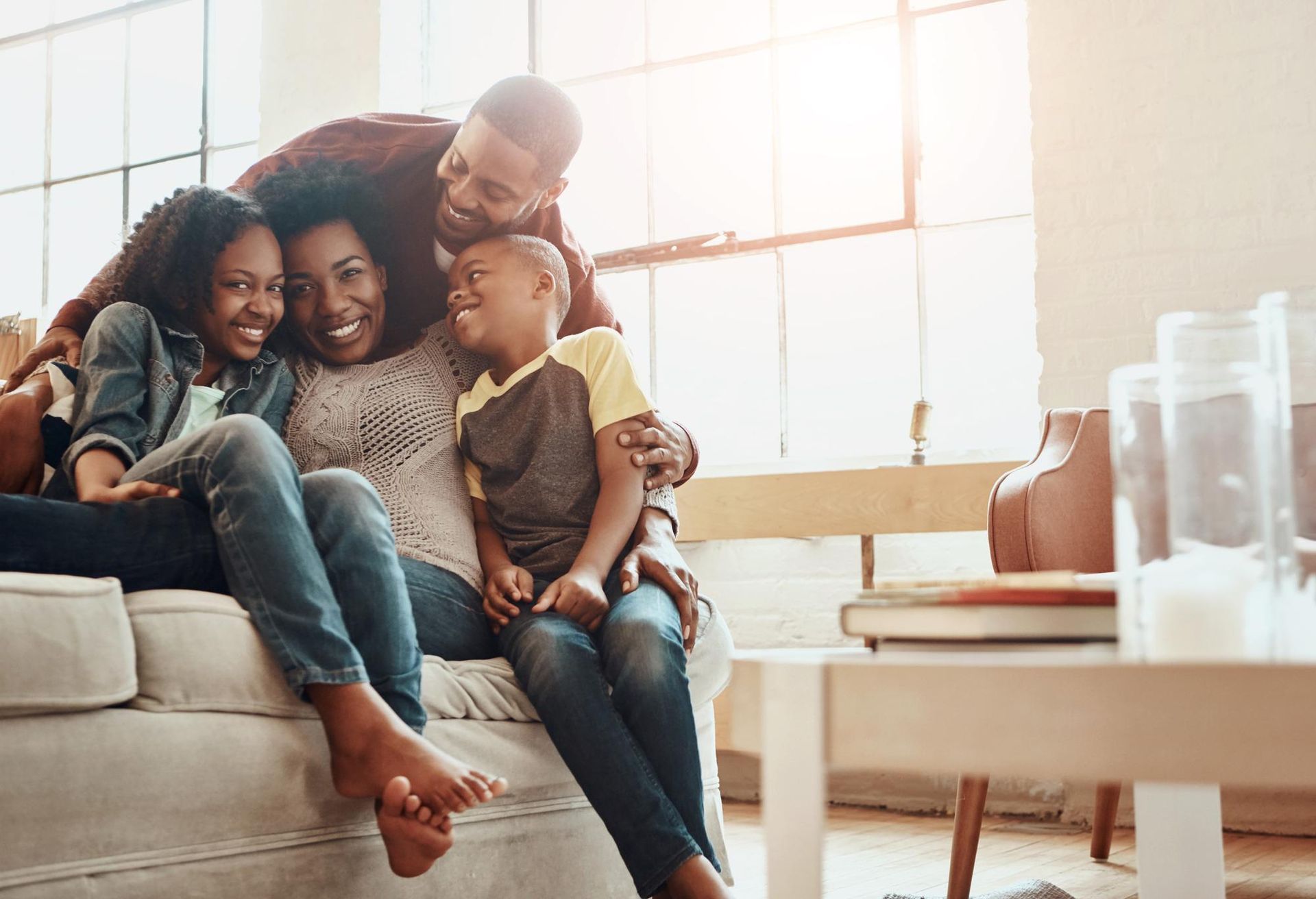A family of four smiles while sitting together on a couch in a bright, sunlit room.