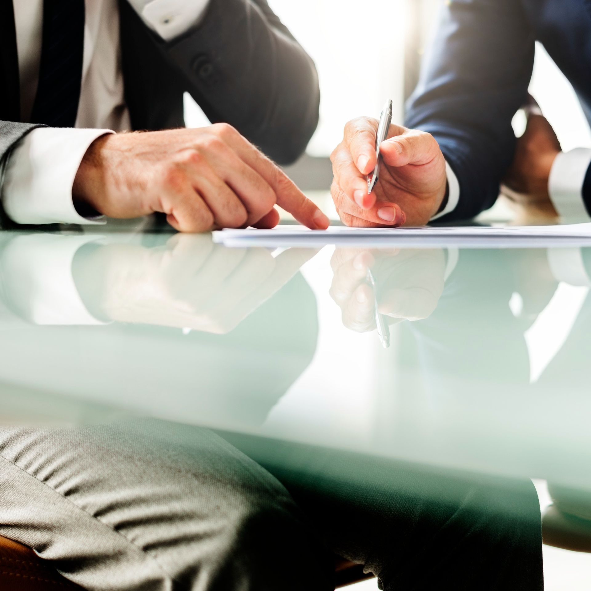 Two people in professional suits sit at a glass table, reviewing a document together.
