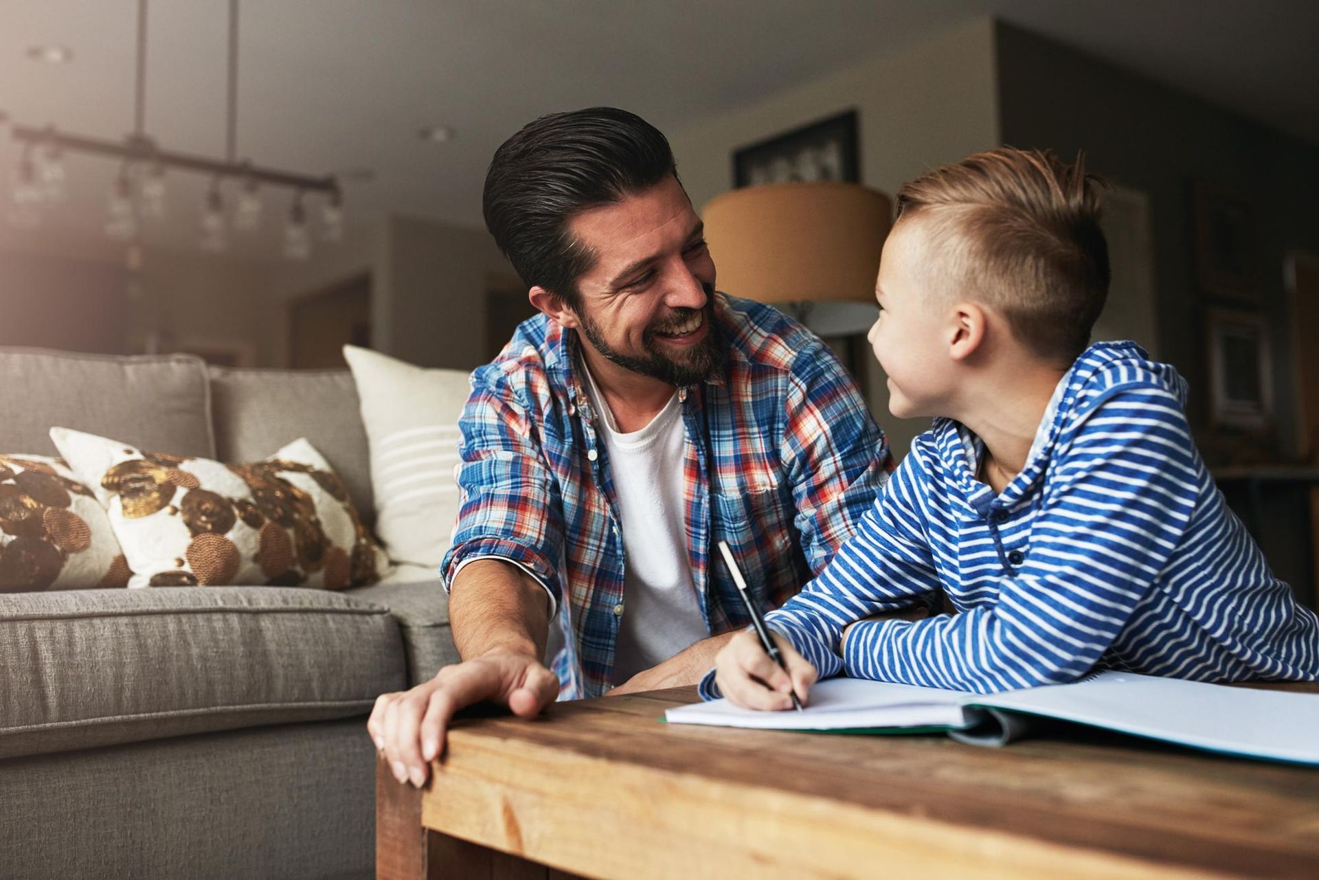 A smiling adult and a child writing in a notebook together on a living room coffee table.