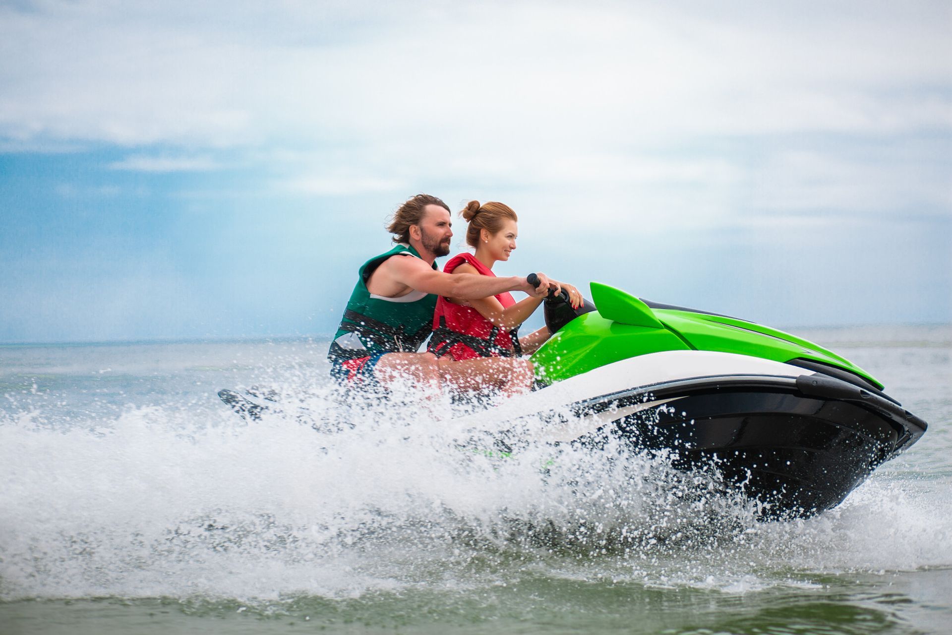 A couple wearing life vests riding a green and white jet ski across the ocean, creating a large splash.