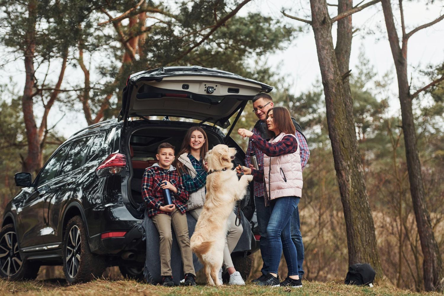 A family stands with their golden retriever near an open car trunk in a wooded area during a daytime outing.