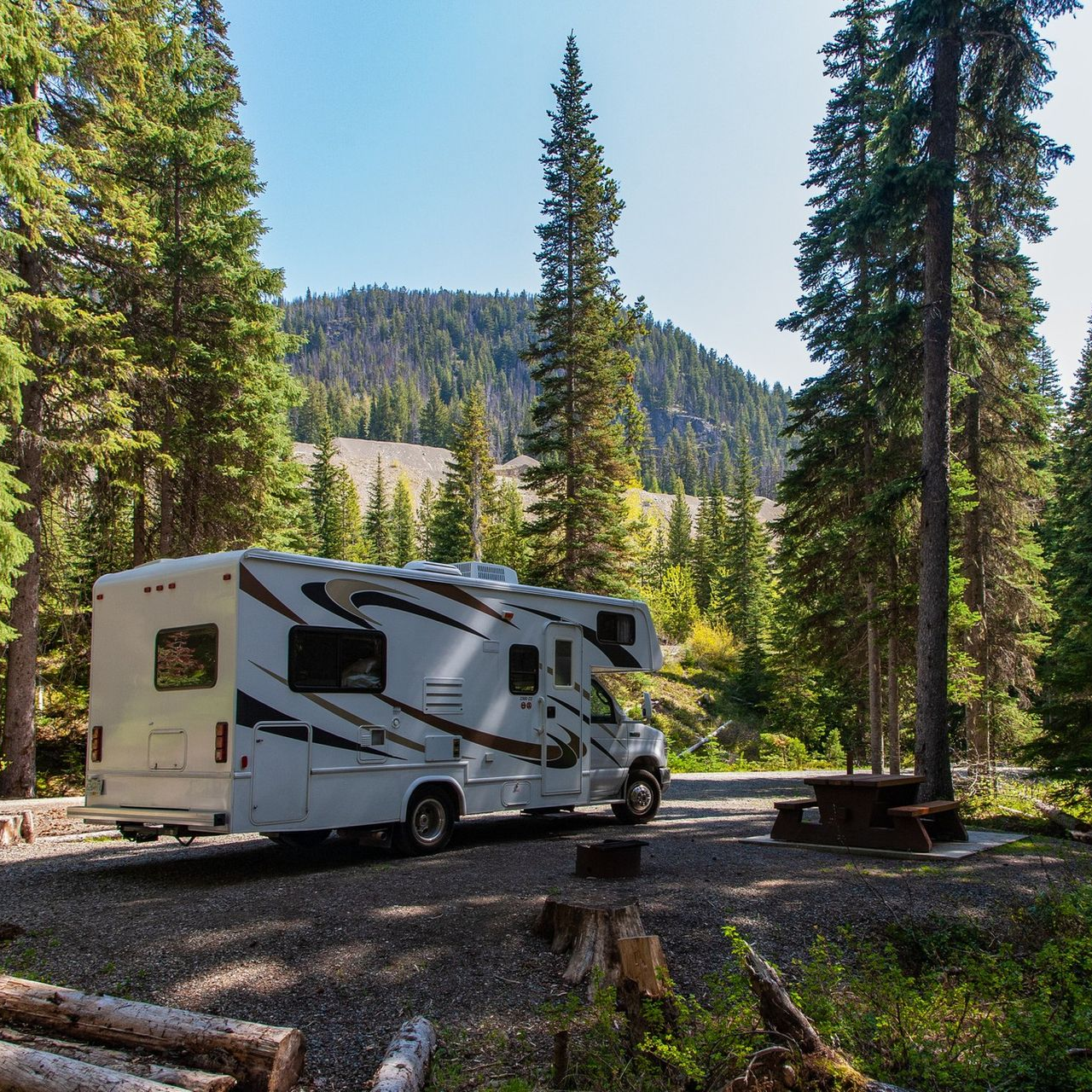 A white recreational vehicle is parked on a gravel campsite surrounded by a dense forest under a clear blue sky.