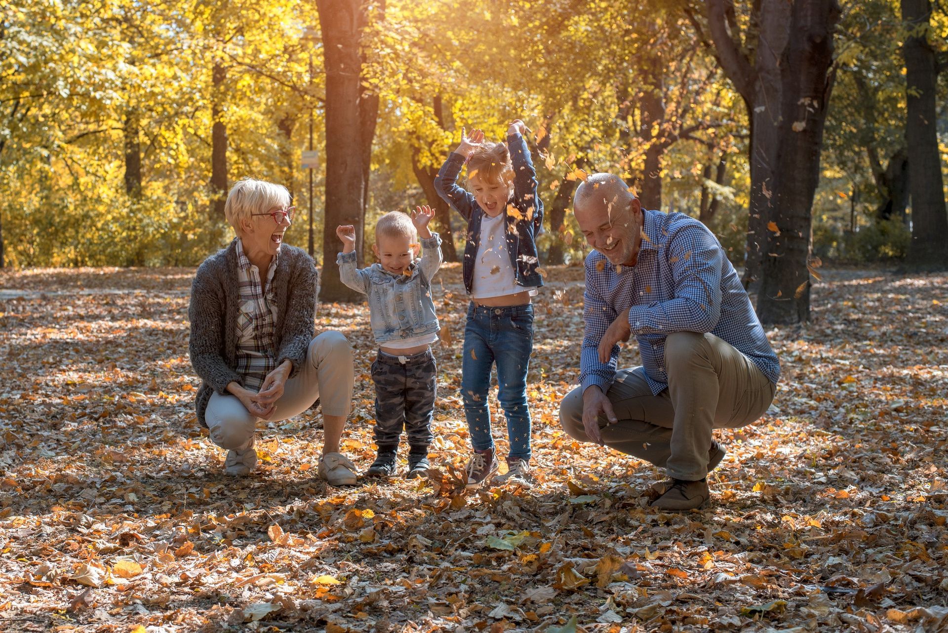Two adults and two children crouch and stand in a sunny, autumn-colored park, tossing dried leaves into the air.