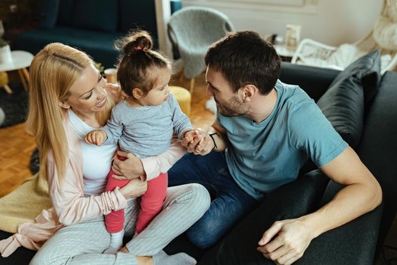 A young family sits on a sofa in a living room, smiling and interacting with their toddler.
