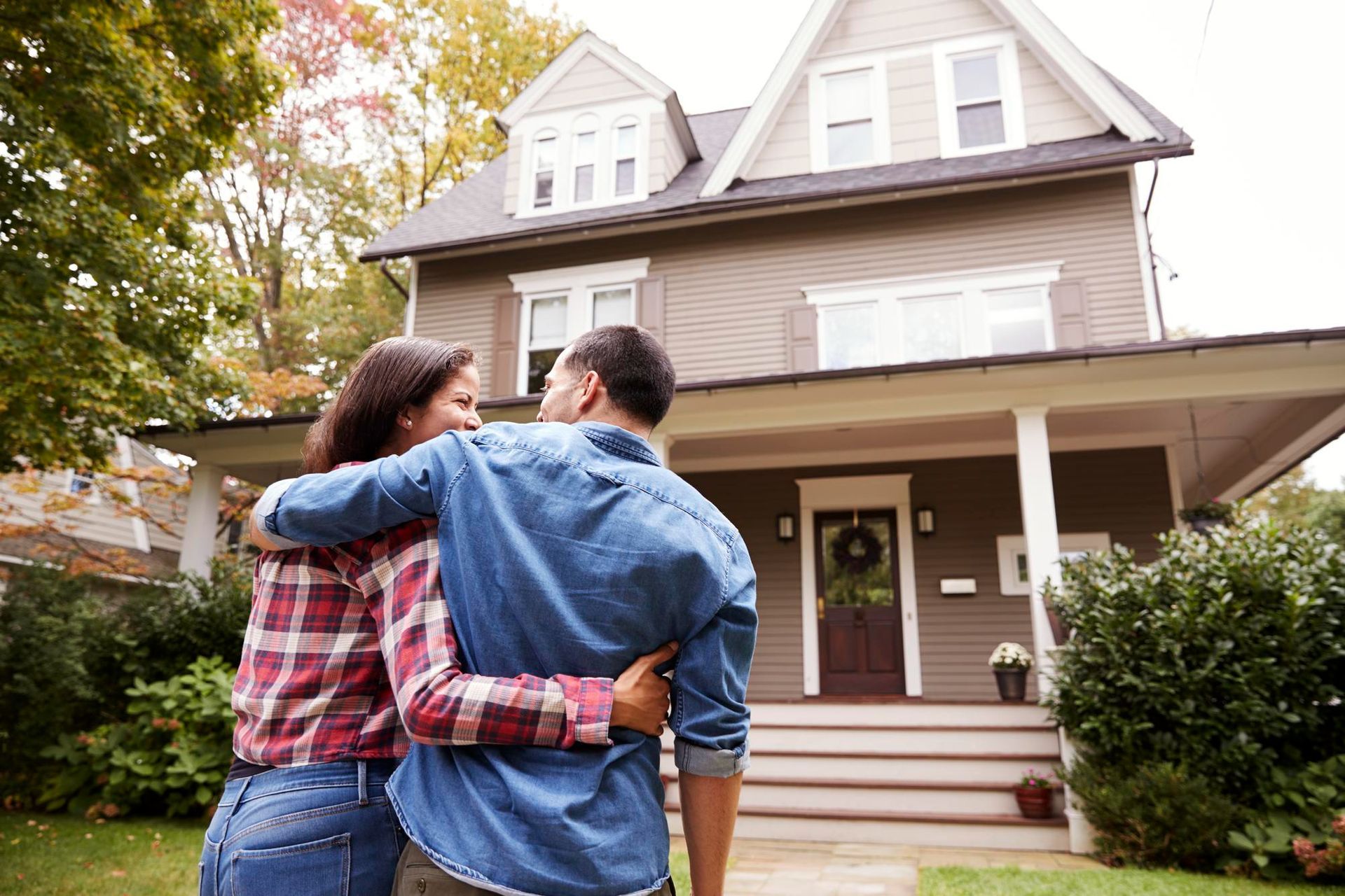 A couple in casual clothing stands with their arms around each other, looking at a two-story suburban home.