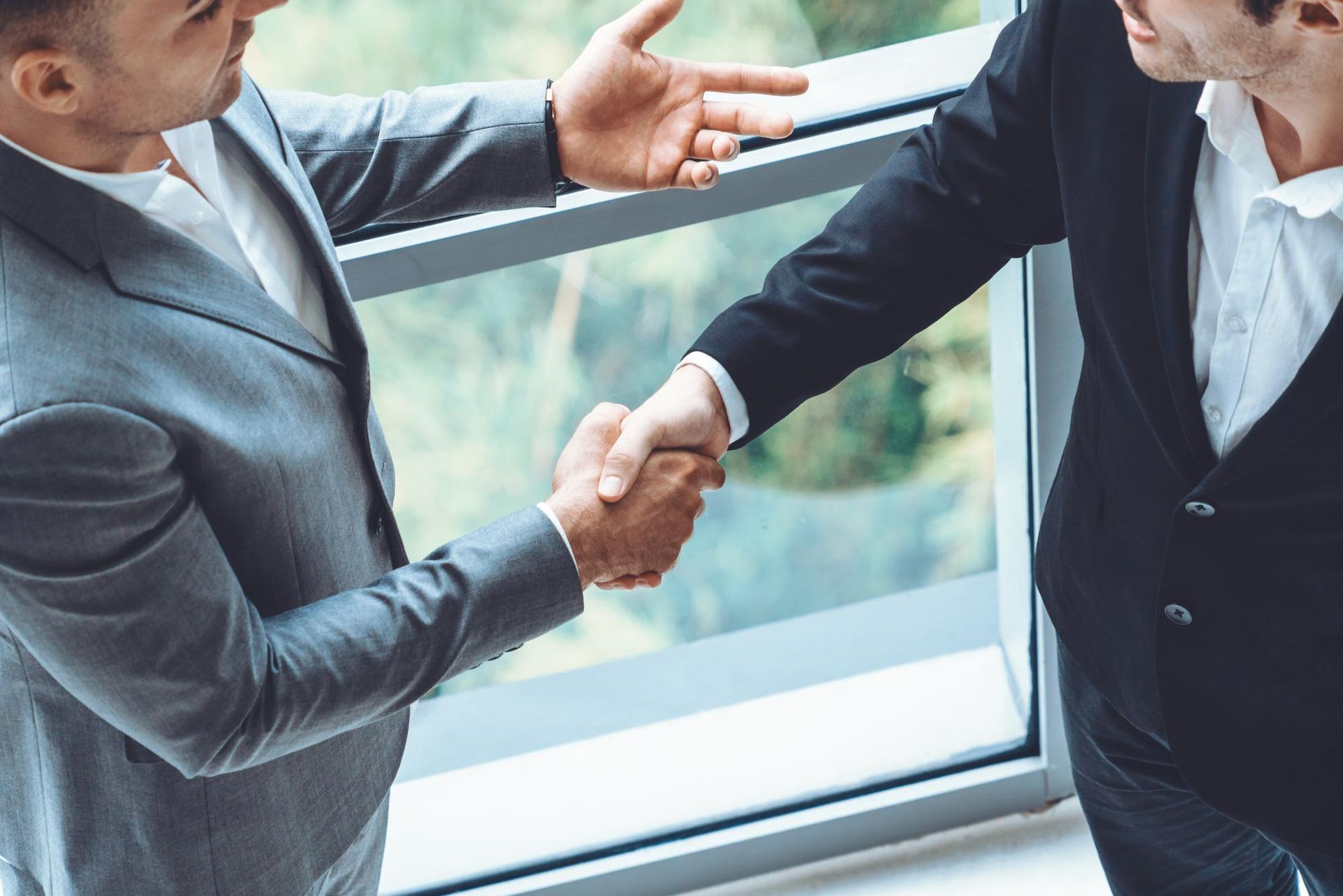 Two business people in suits shake hands near a large window, with one gesturing while they converse.