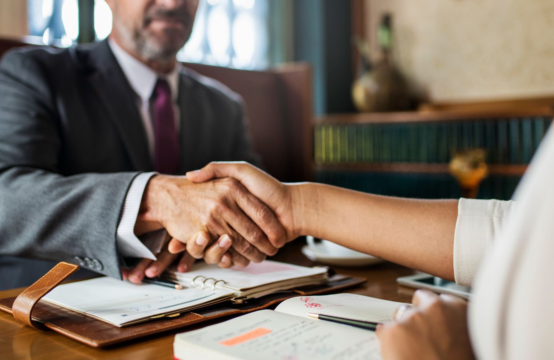 Two people shake hands over an open notebook and documents on a desk in a professional office setting.