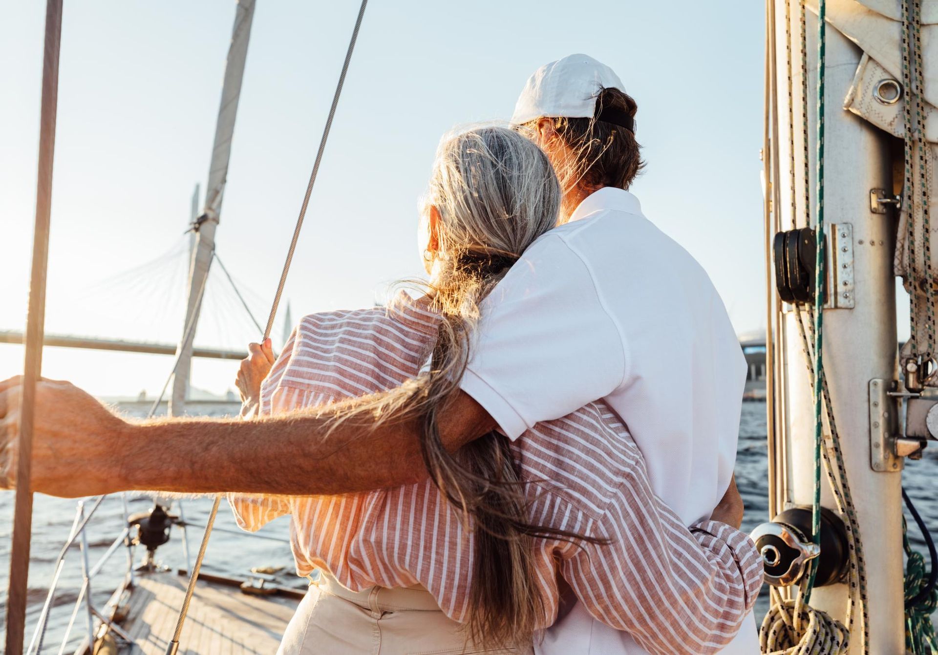 Two people in light clothing embrace while standing on a sailboat at sunset, looking out toward the water.
