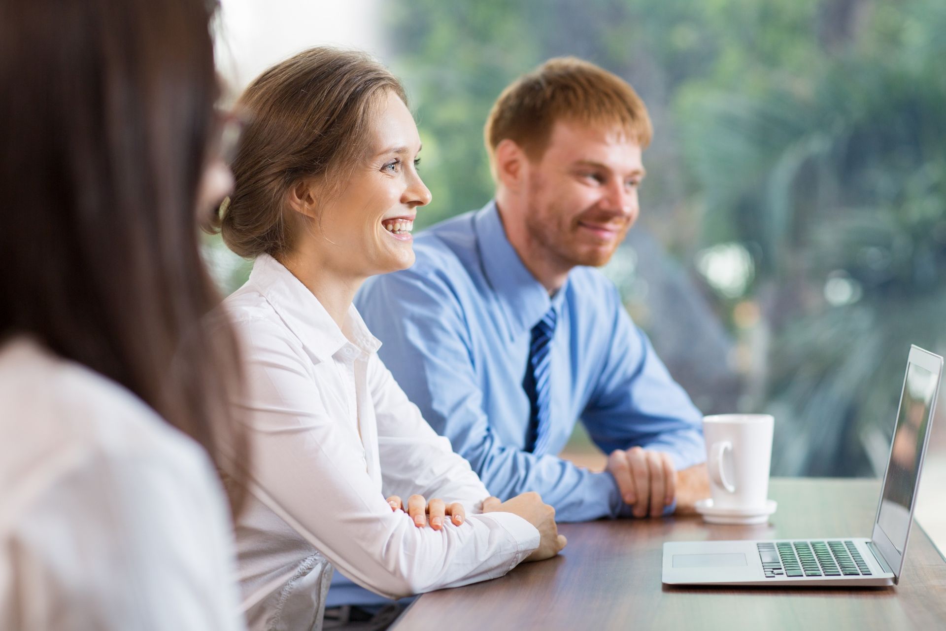 A group of colleagues in professional attire sitting at a desk with a laptop and coffee, looking at a screen and smiling.