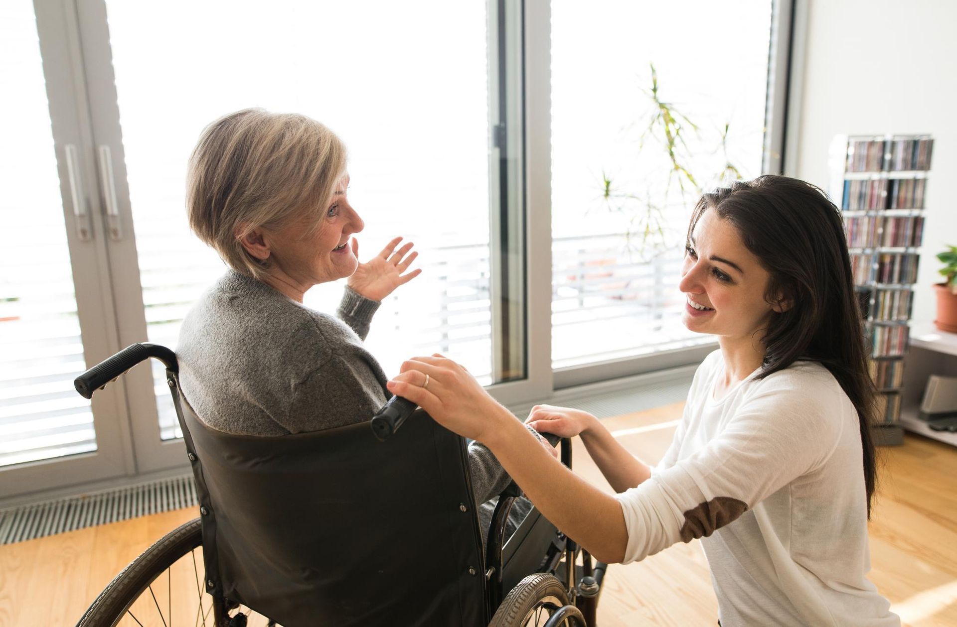 A woman in a wheelchair speaks with a caregiver who has a hand on the wheelchair handle in a bright, sunlit room.