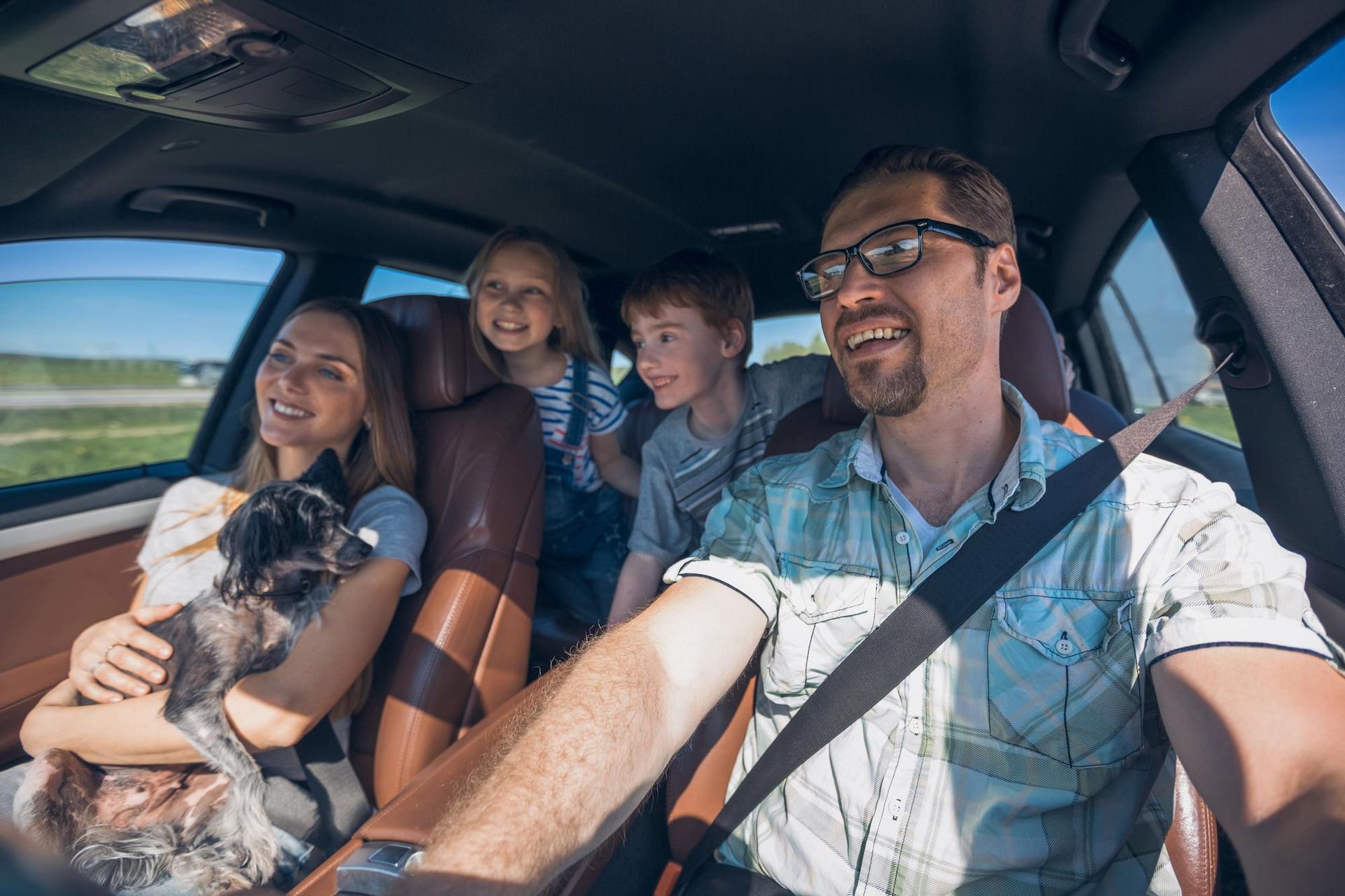 A smiling family drives in a car; the driver is at the wheel, while a woman holds a small dog in the passenger seat.