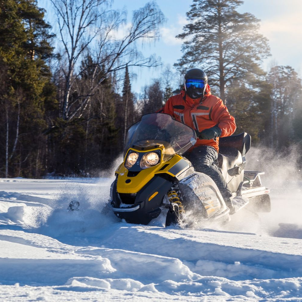 A person in an orange jacket rides a yellow and black snowmobile through deep snow in a sunlit forest.