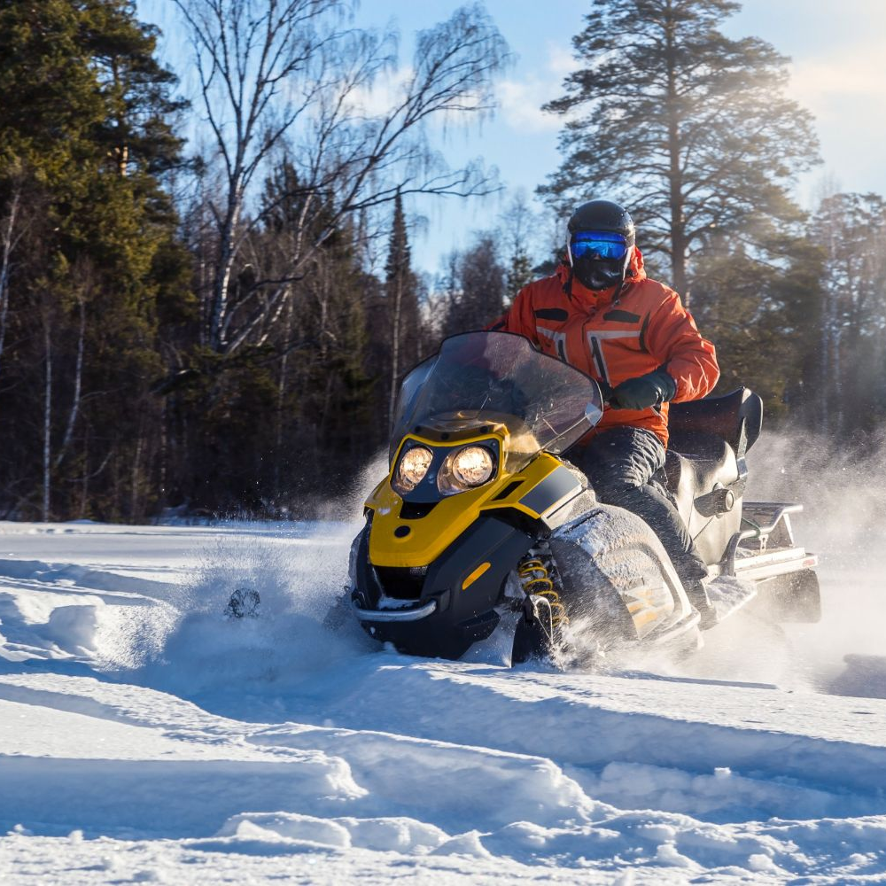 A person in an orange jacket rides a yellow and black snowmobile through snowy woods on a bright, sunny day.