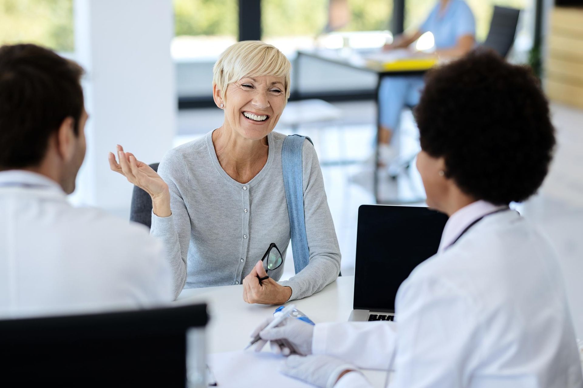 A smiling patient in a consultation room gestures while talking to two medical professionals wearing white coats.