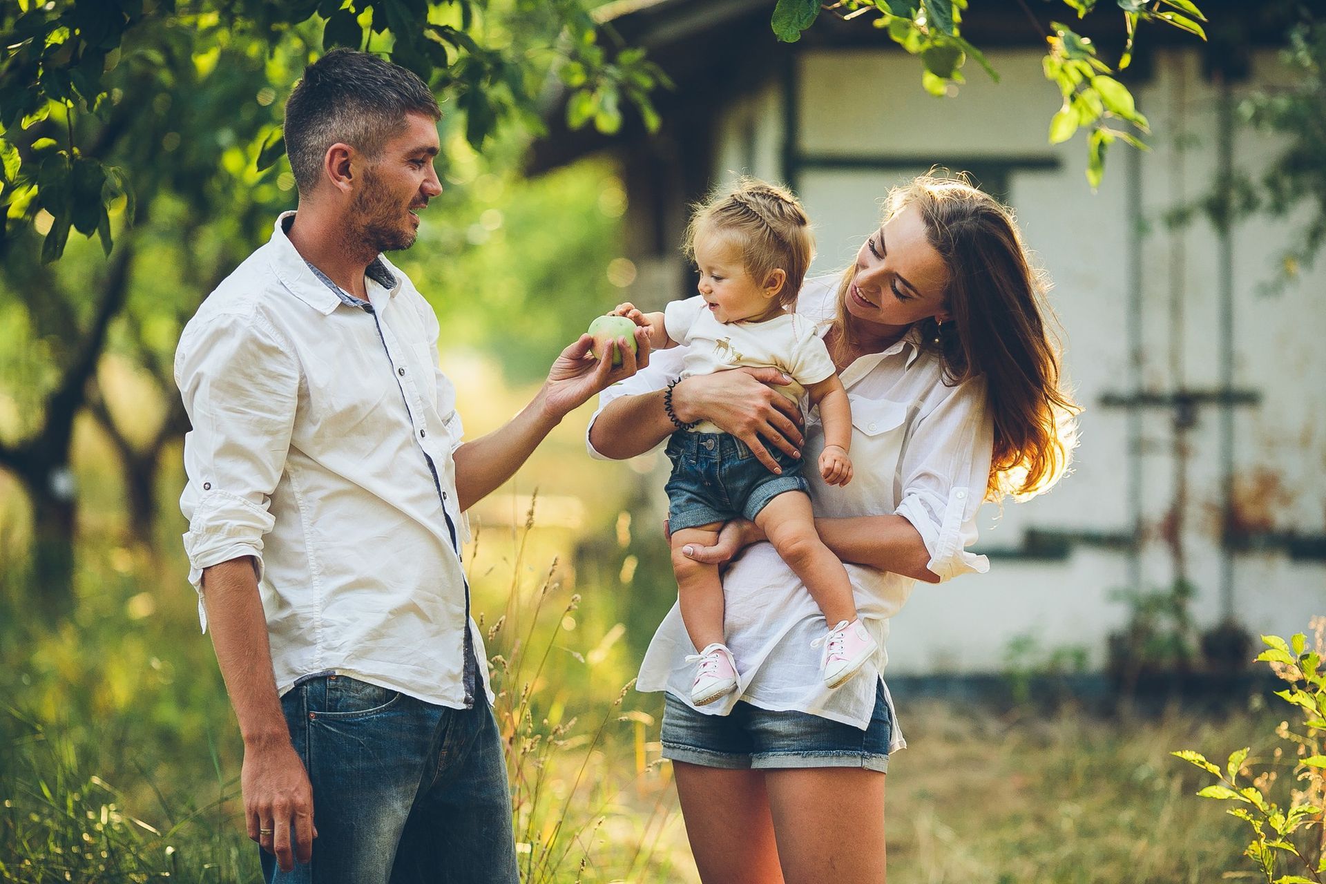 A family stands in a sunny garden; a man offers an apple to a toddler held by a woman.