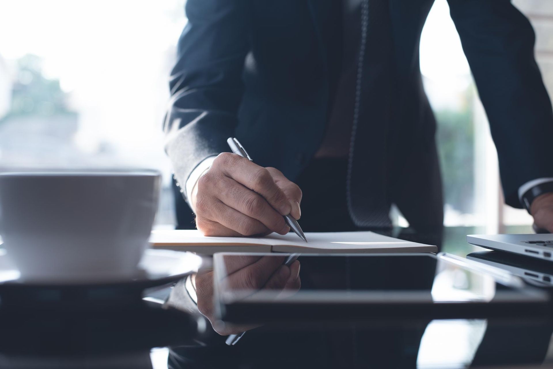 A person in a dark suit writing in a notebook on a reflective office desk with a coffee cup nearby.
