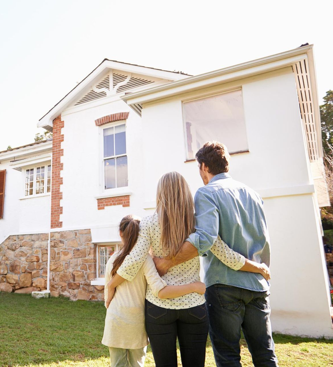 A family stands with their arms around each other, looking up at their white, two-story house with stone foundation accents.