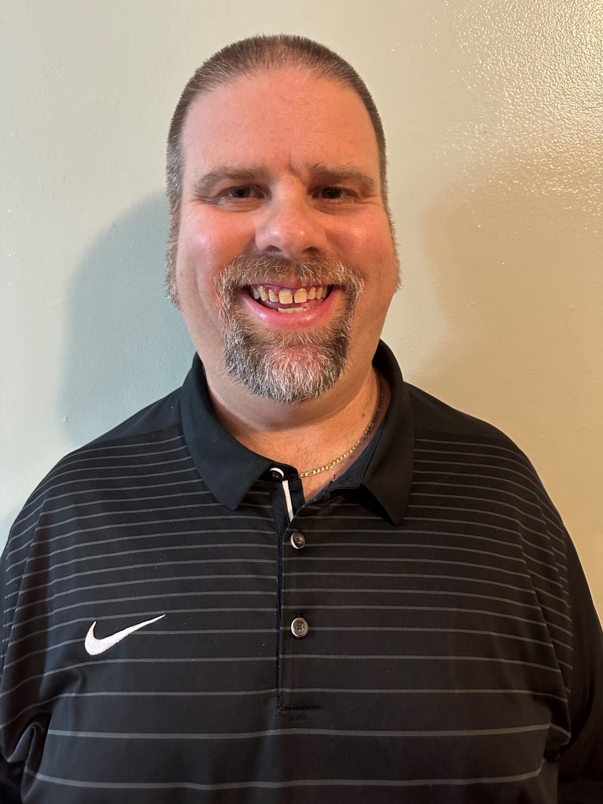 A person with a goatee smiles at the camera, wearing a black collared shirt with thin horizontal stripes.