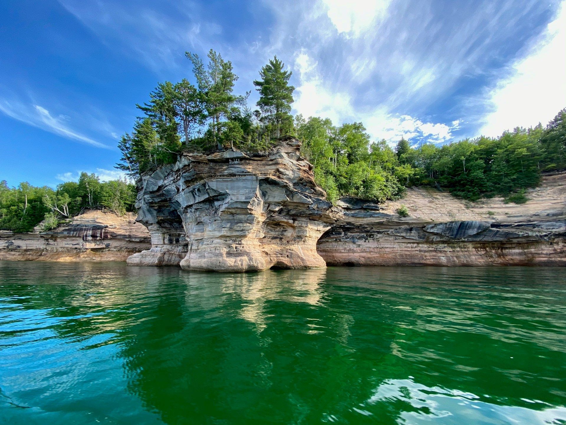 A sandstone cliff topped with green trees rising out of the clear, vibrant green water of Lake Superior.