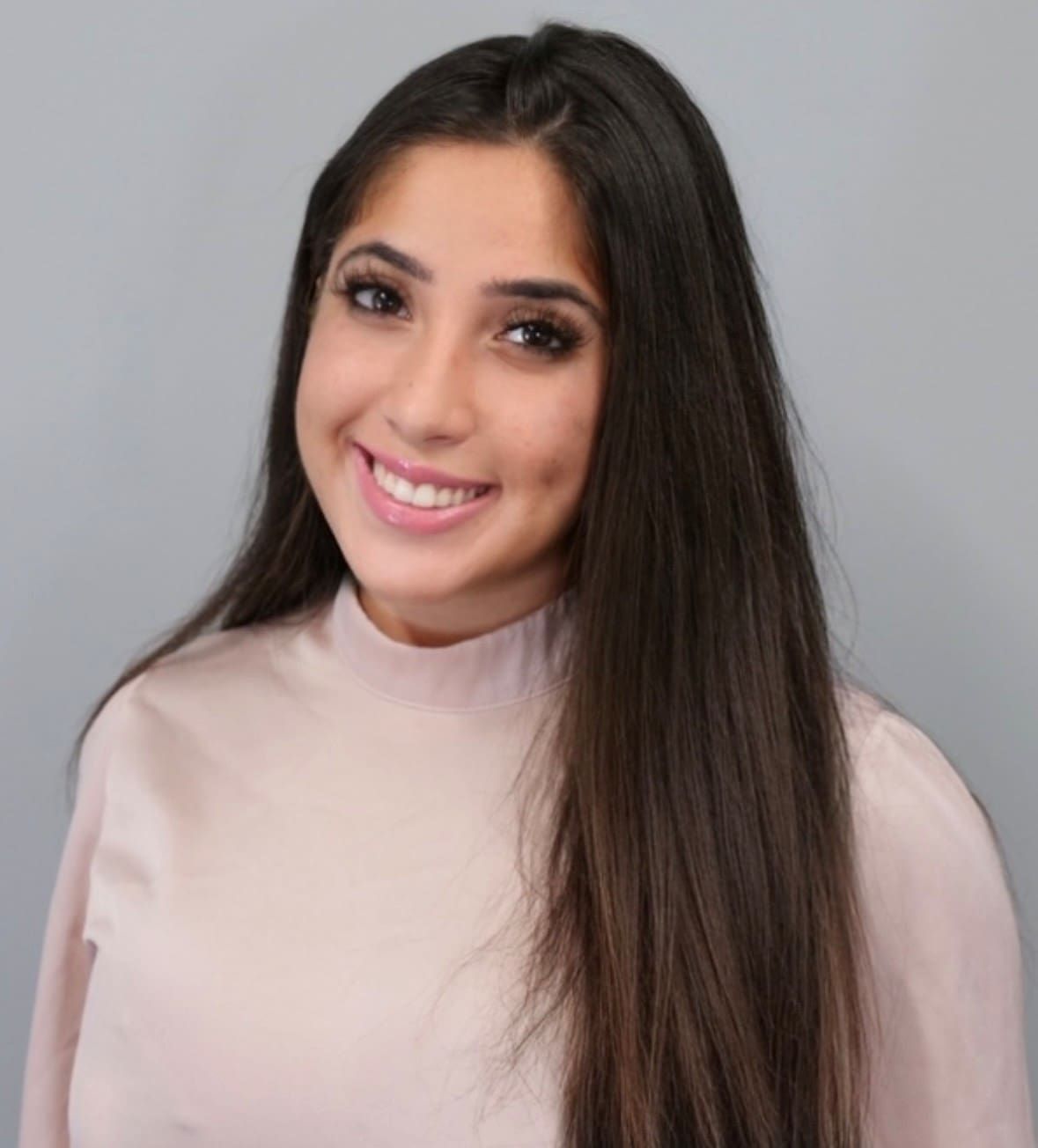 A smiling person with long brown hair wearing a light pink high-neck top against a plain gray background.
