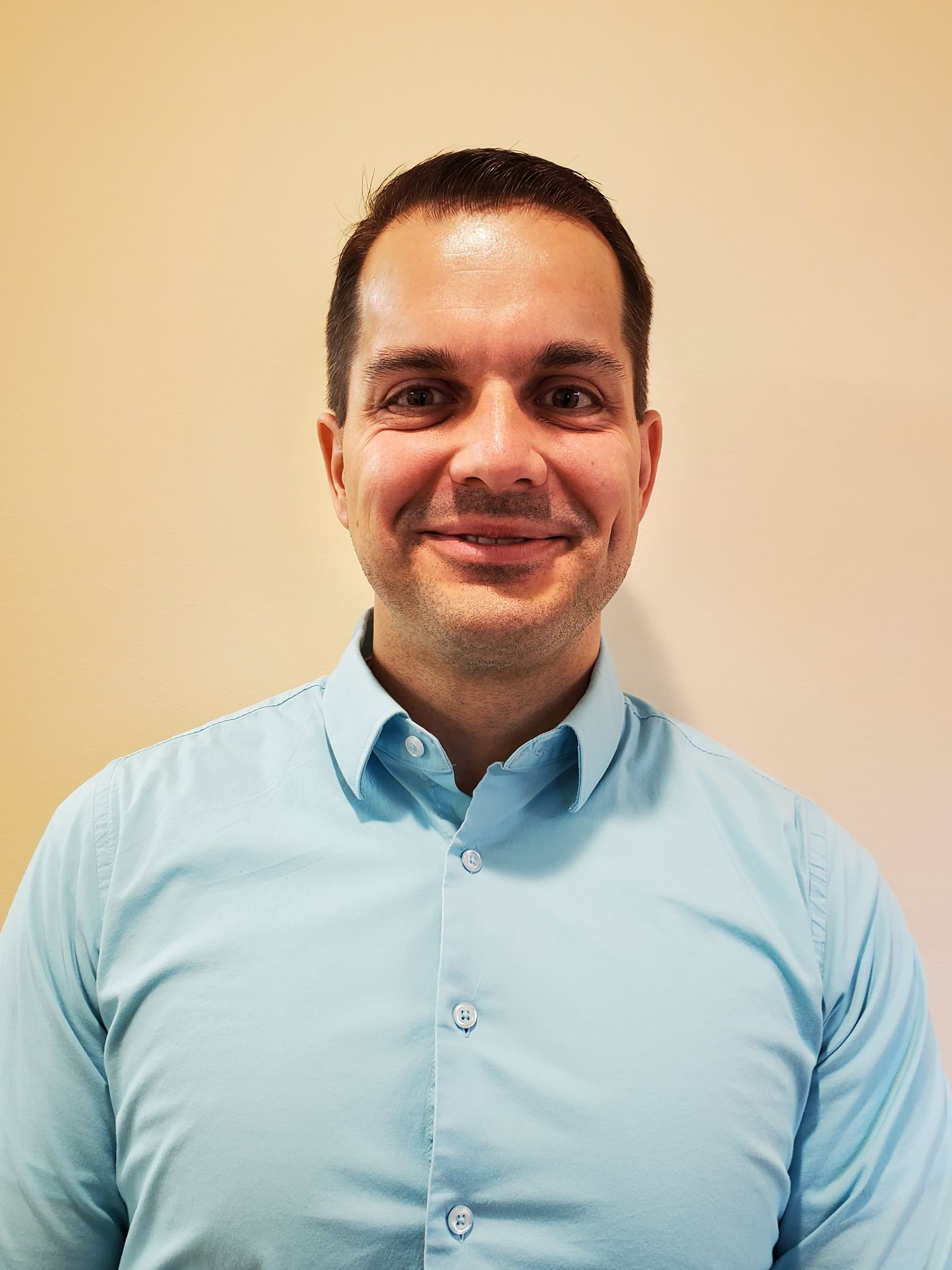 A person with short dark hair and a light blue button-down shirt smiling against a light tan background.