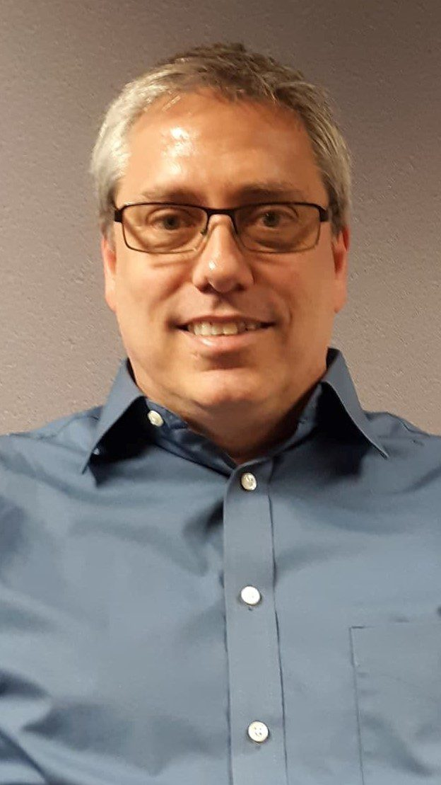 A smiling person with glasses and graying hair, wearing a blue button-down shirt against a plain, light-colored background.