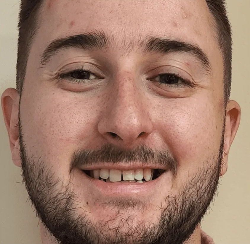 A close-up portrait of a person with a short beard, light complexion, and brown eyes, smiling directly at the camera.