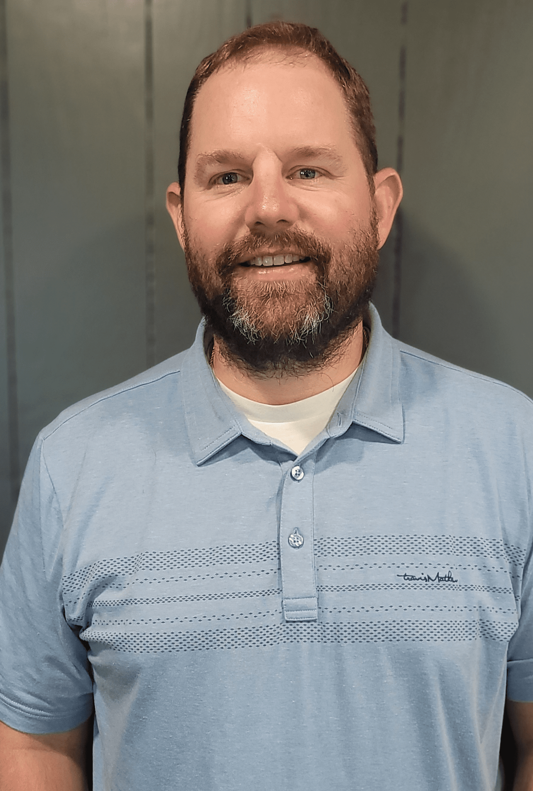 A smiling person with a beard, wearing a light blue polo shirt over a white undershirt, stands against a gray wall.
