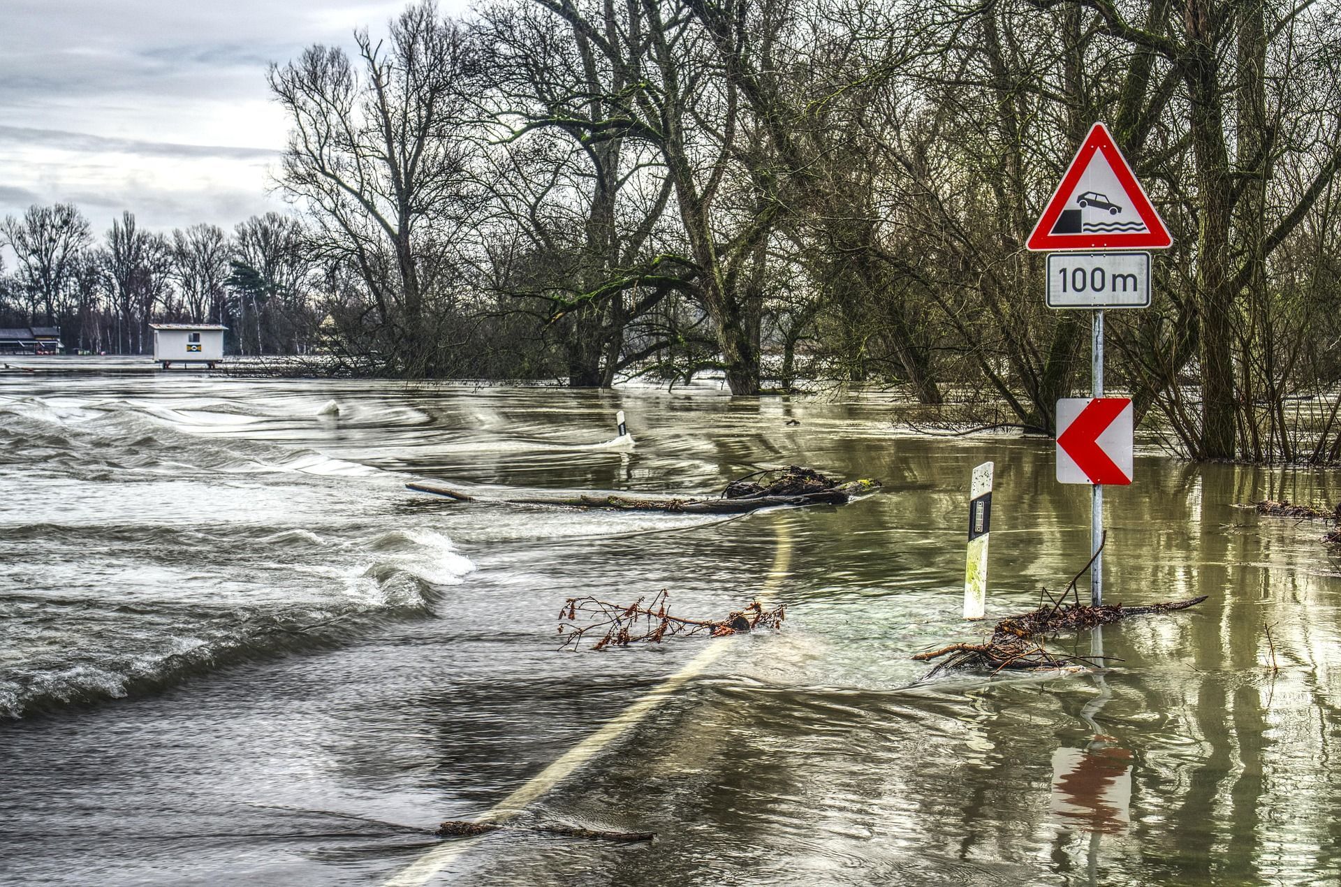 Floodwaters cover a road near trees, with a warning sign indicating a quay or drop-off in 100 meters.
