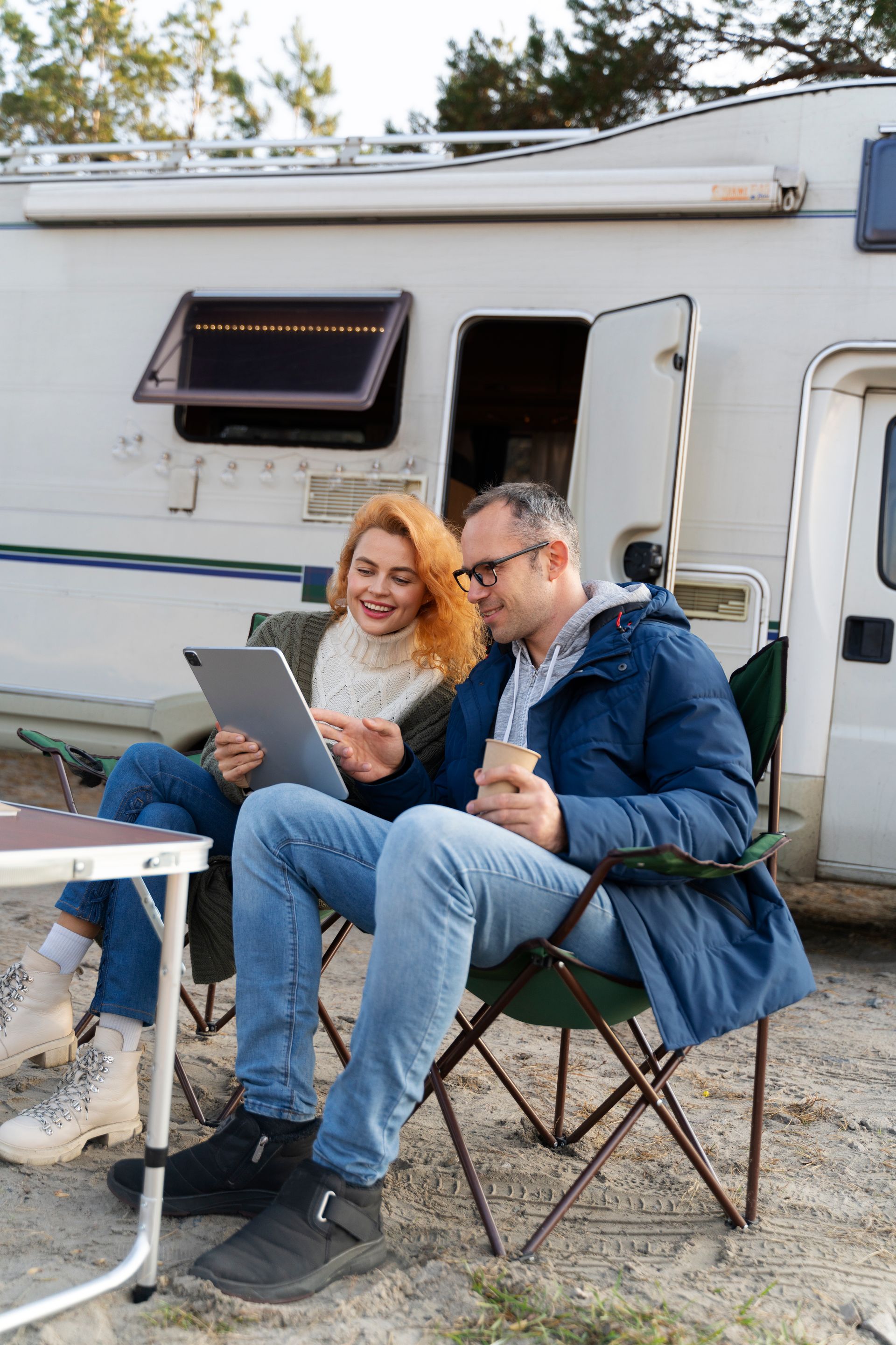 A couple sits in camping chairs outside a camper van, smiling while looking at a tablet together.