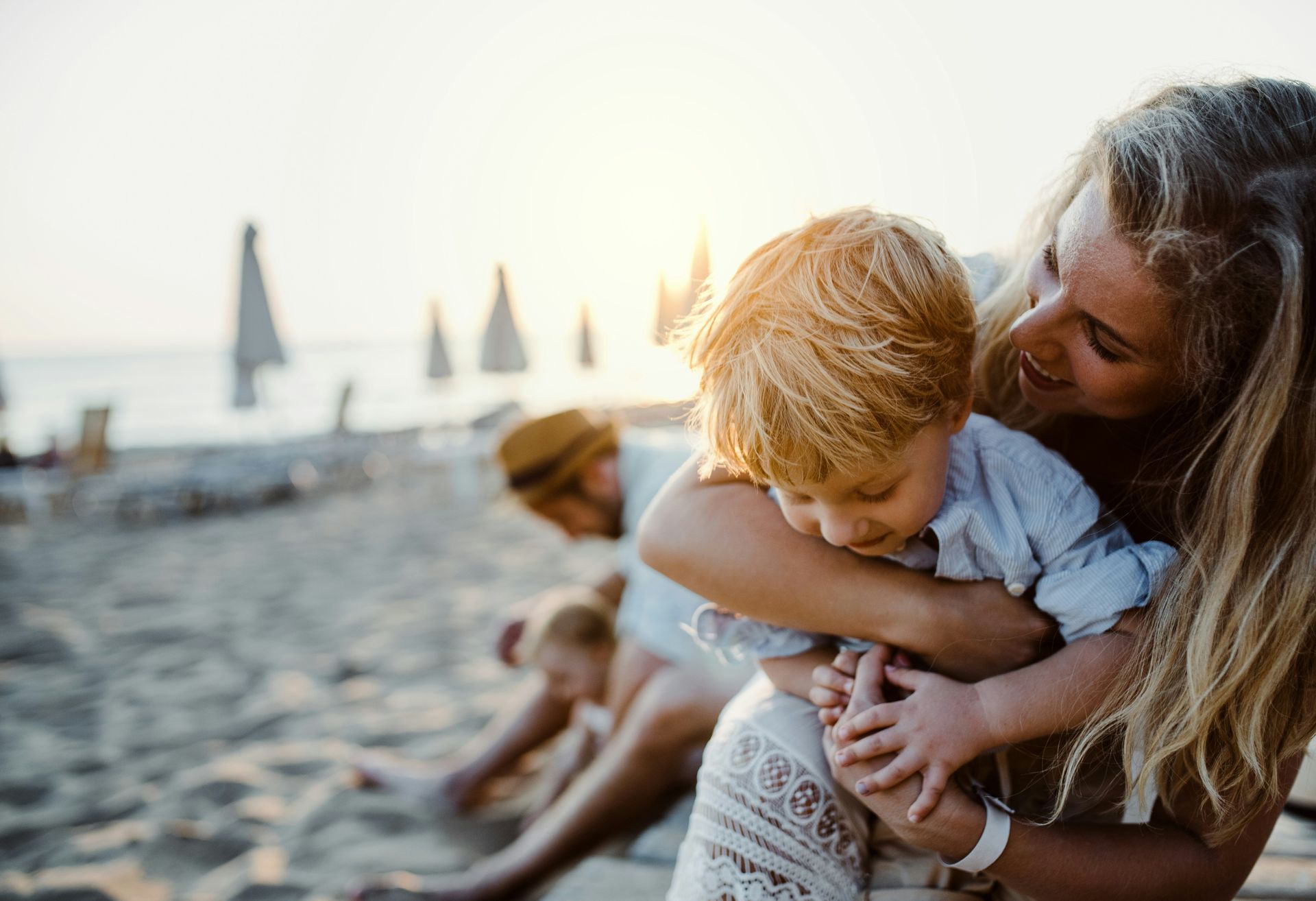 A person embraces a young child on a sandy beach at sunset, with another person sitting nearby in the background.