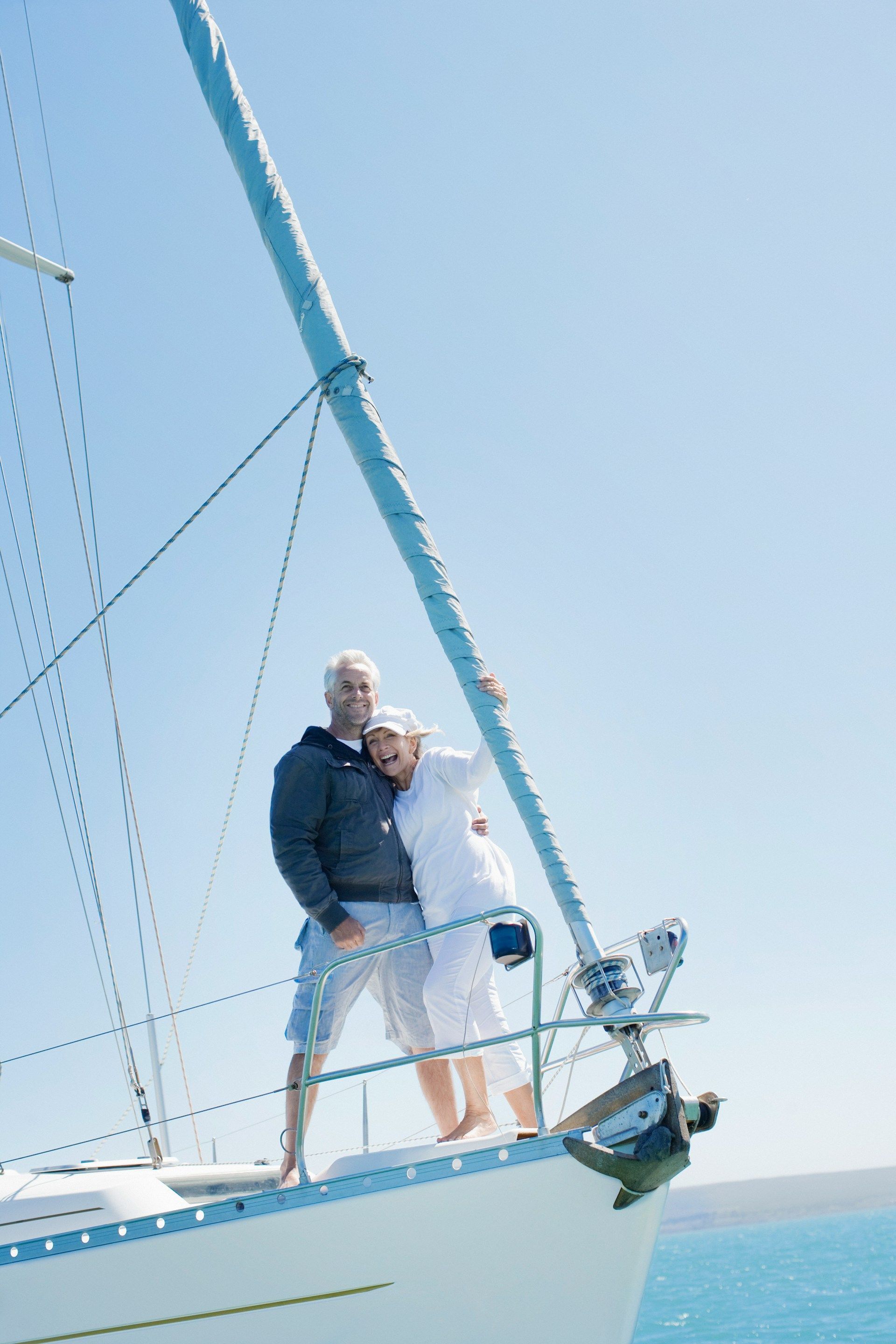 An older couple standing together on the deck of a sailboat on the water under a clear blue sky.