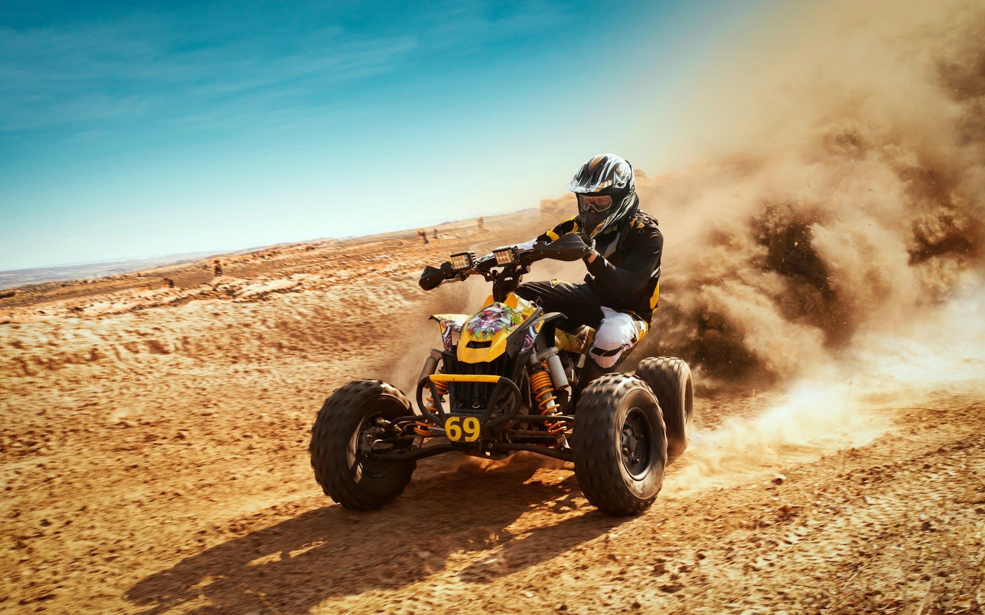 A person in safety gear rides a yellow and black ATV, kicking up a large cloud of dust in a desert landscape.
