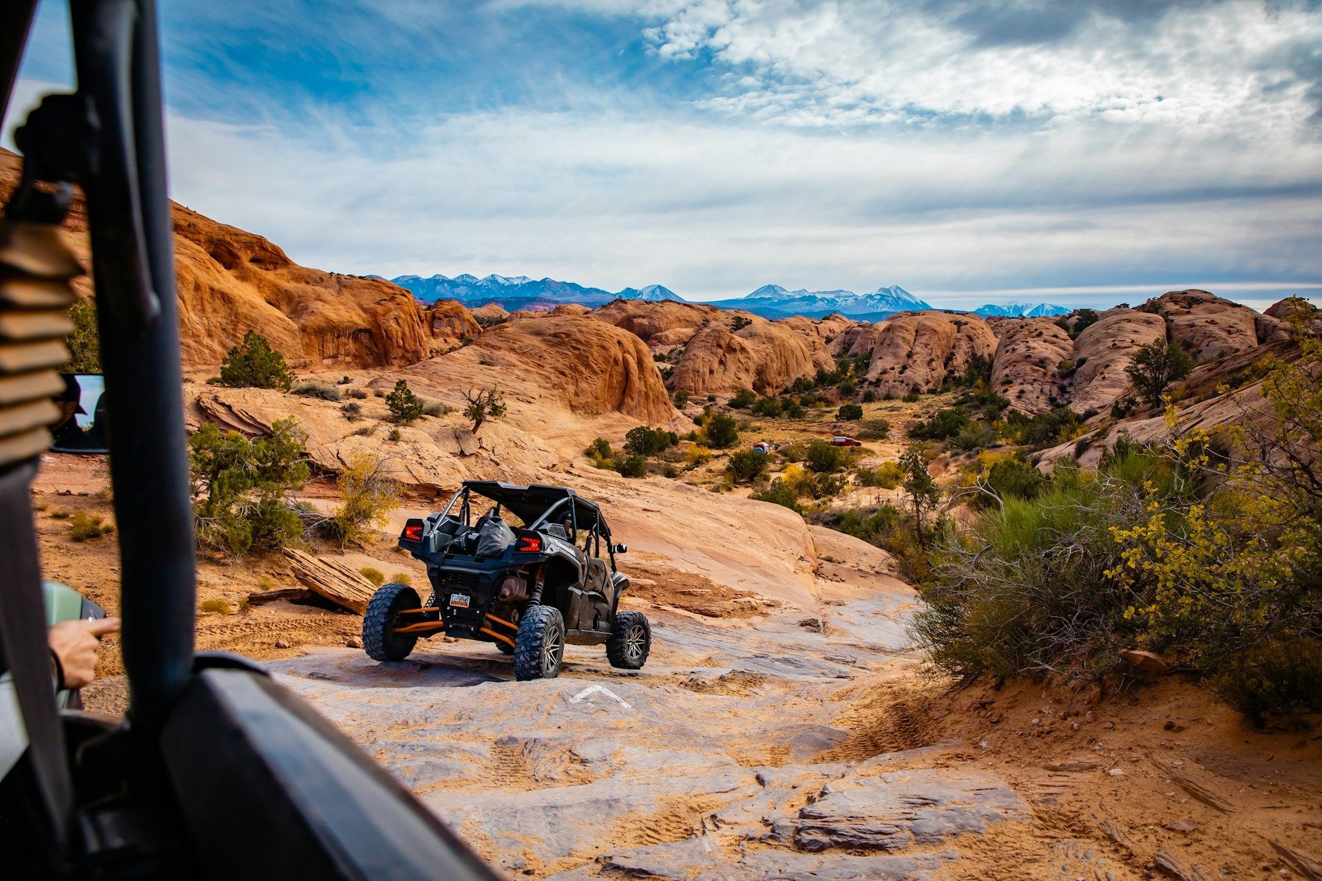 A side-by-side utility vehicle drives over rocky terrain in a desert landscape with distant mountains under a cloudy sky.