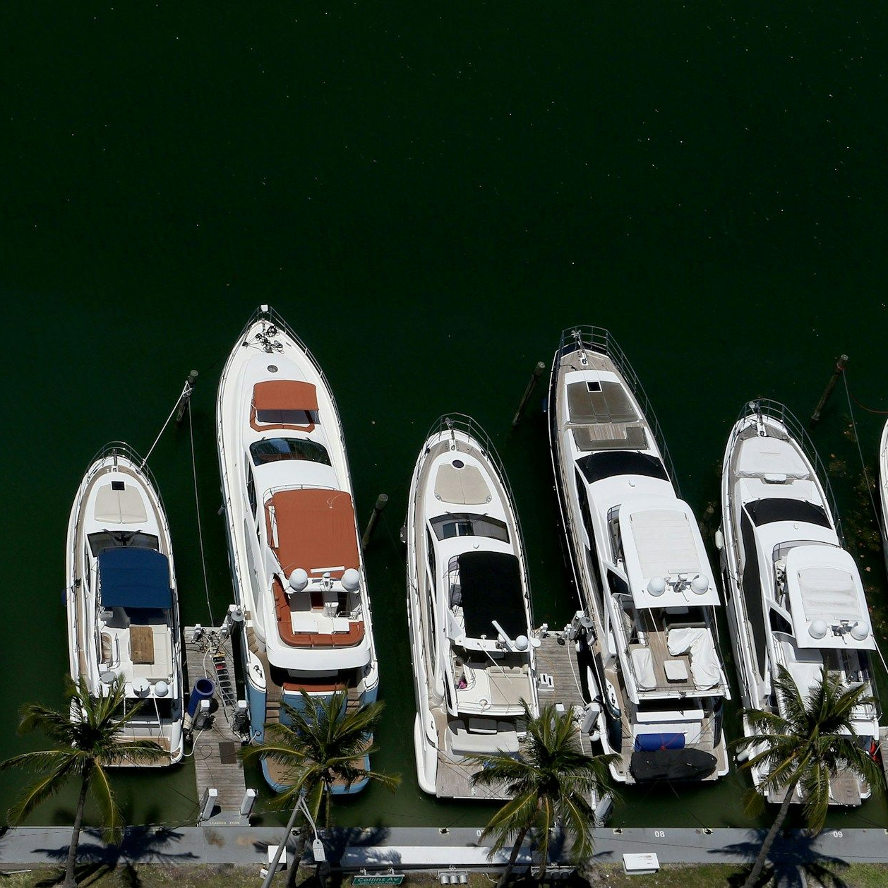 Aerial view of five white yachts docked side-by-side at a pier, with palm trees visible along the edge.