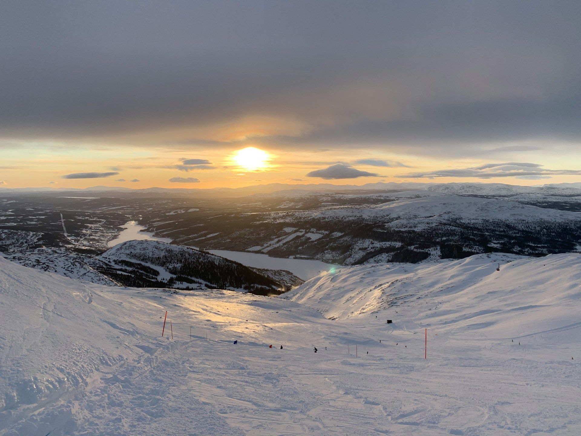 A golden sunset glows over a vast, snow-covered mountain landscape and a valley lake viewed from a ski slope.