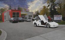 A white Lamborghini with scissor doors open sits in a parking lot in front of a building with red and grey walls.