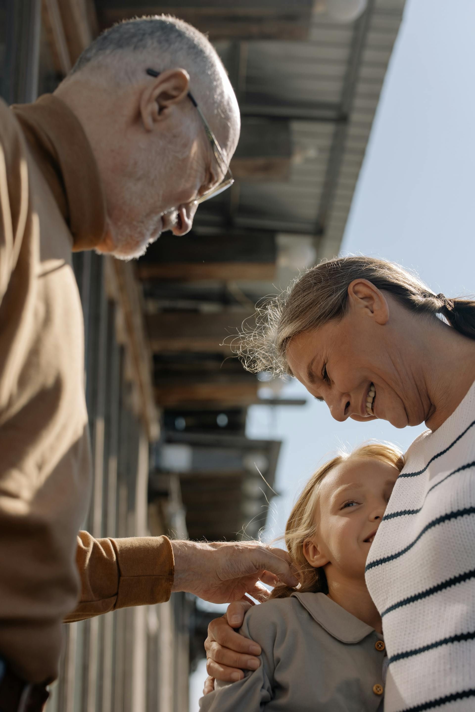 An adult touches the shoulder of a child held by another adult in an outdoor, sunlit setting.
