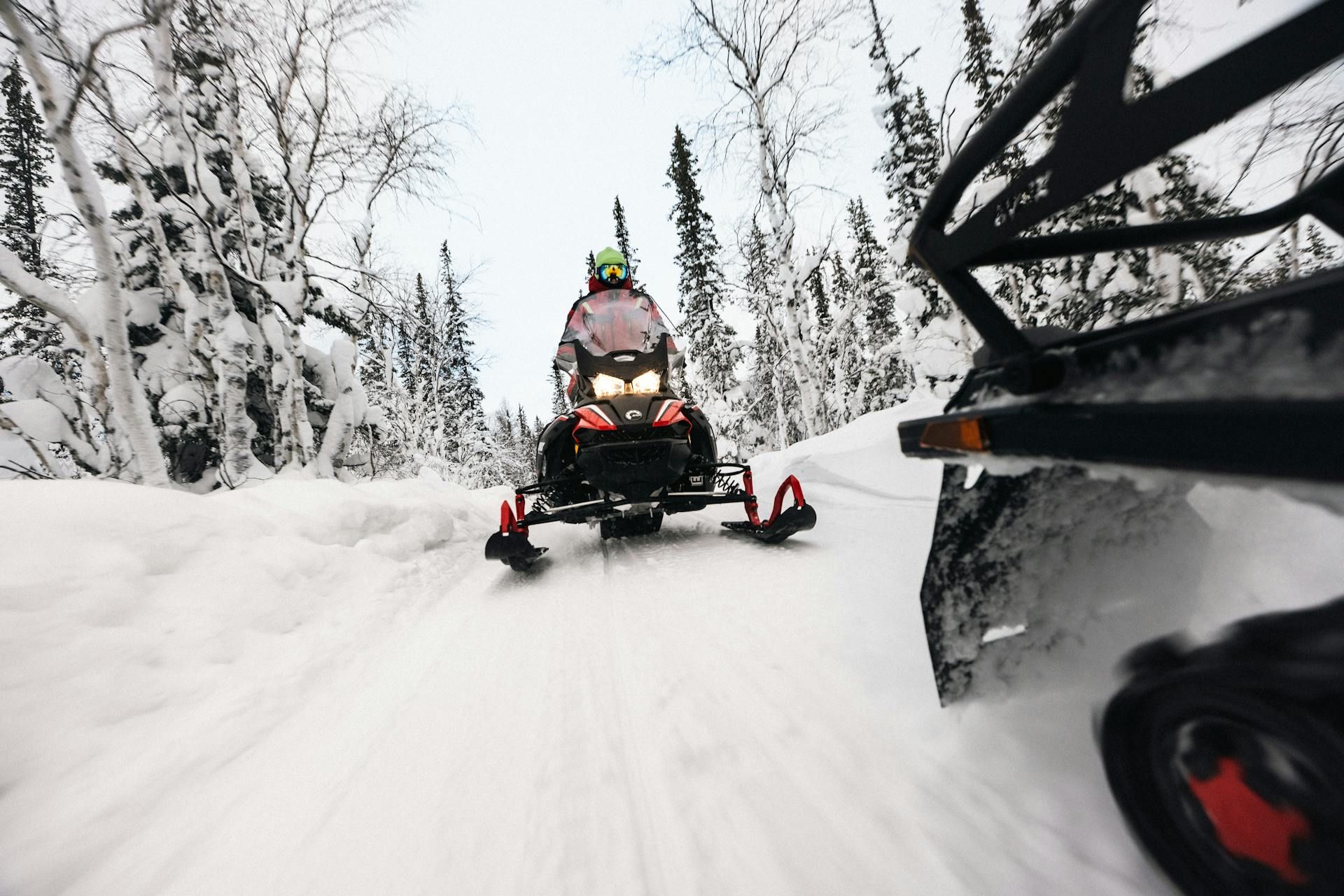 A person on a snowmobile driving toward the camera along a snow-covered trail surrounded by a forest of snow-laden trees.