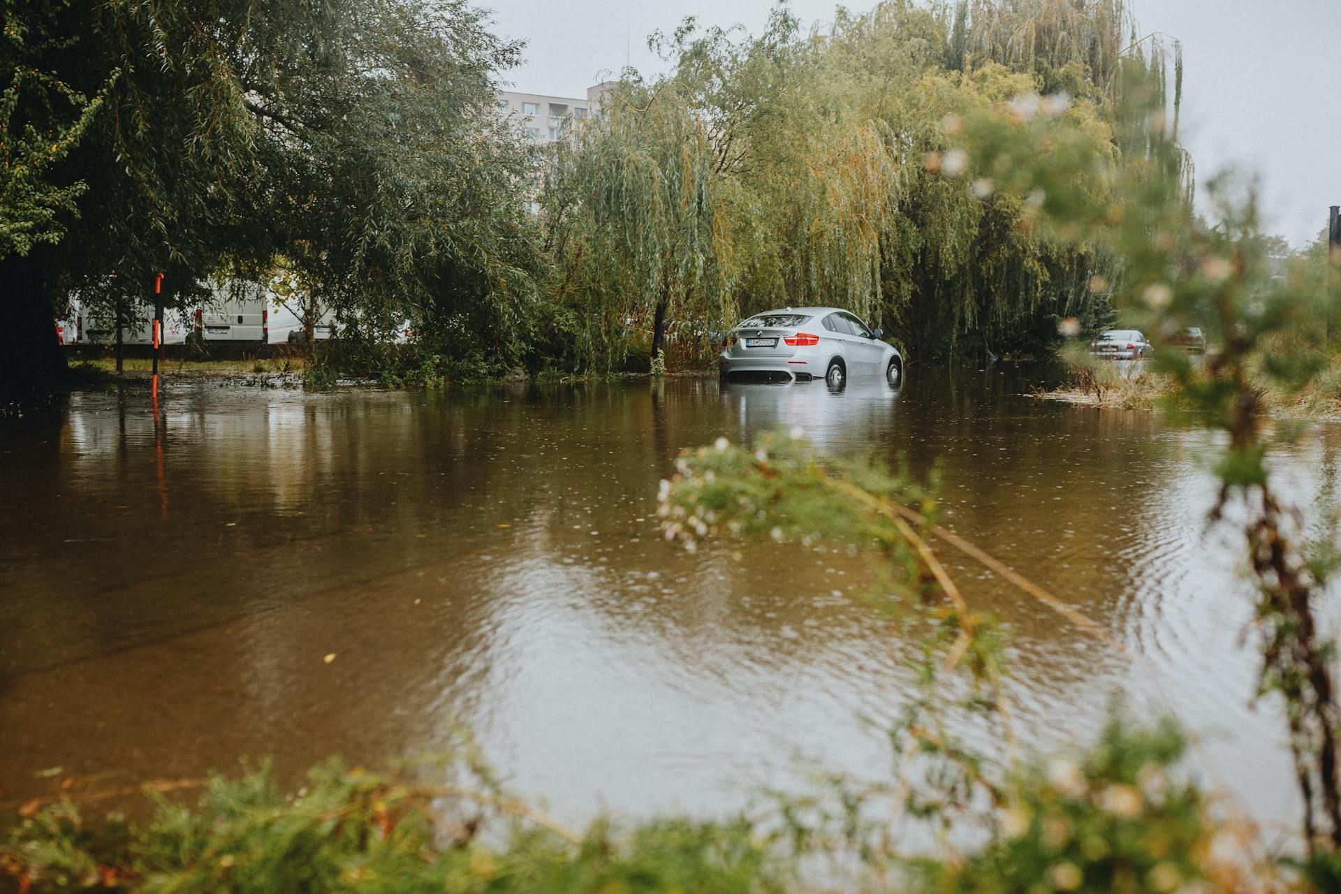 A silver sedan sits partially submerged in a flooded residential street surrounded by trees under a rainy sky.