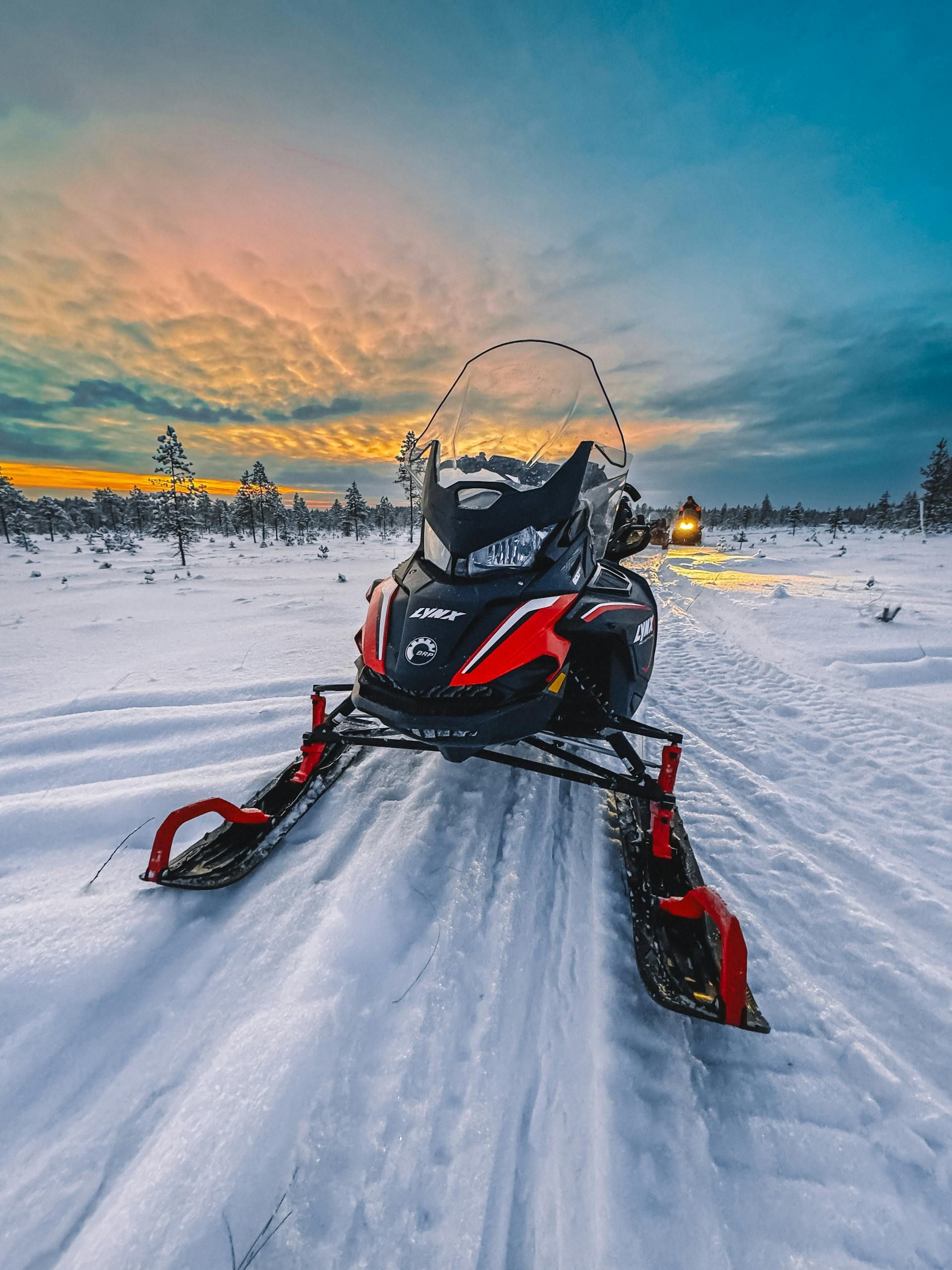 A red and black snowmobile sits on a snow-covered trail at sunset, with another snowmobile visible in the distance.