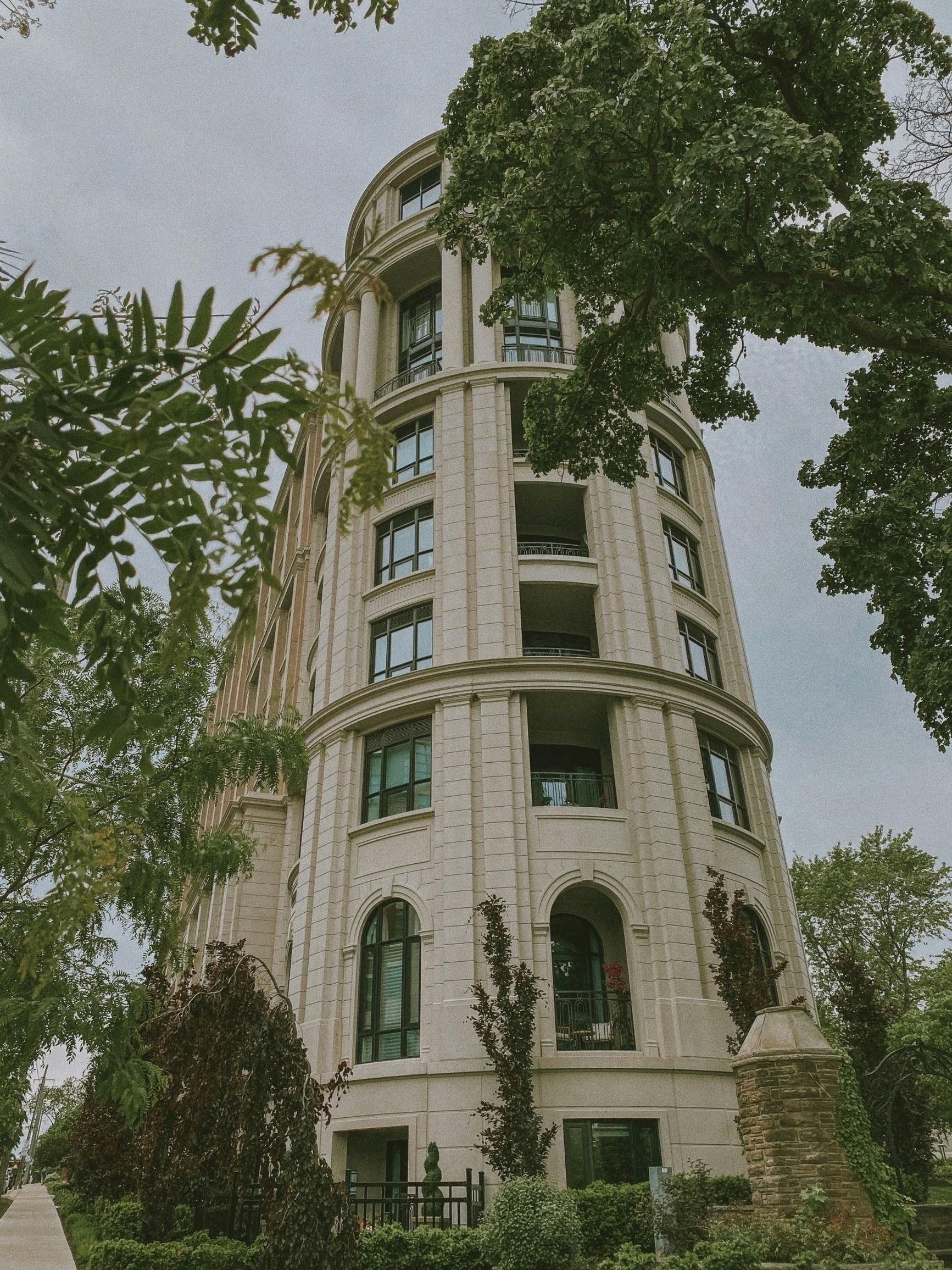 A tall, beige, neoclassical stone building with rounded balconies and arched windows, framed by lush green trees.