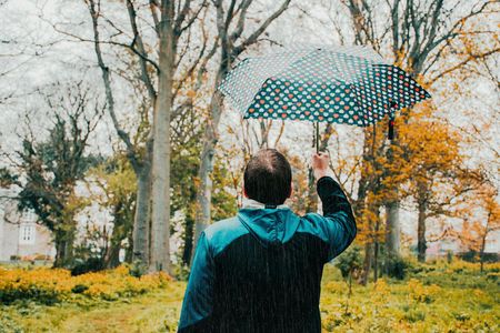 Two people wearing beige trench coats embrace beneath a dark umbrella against a bright, overcast sky.